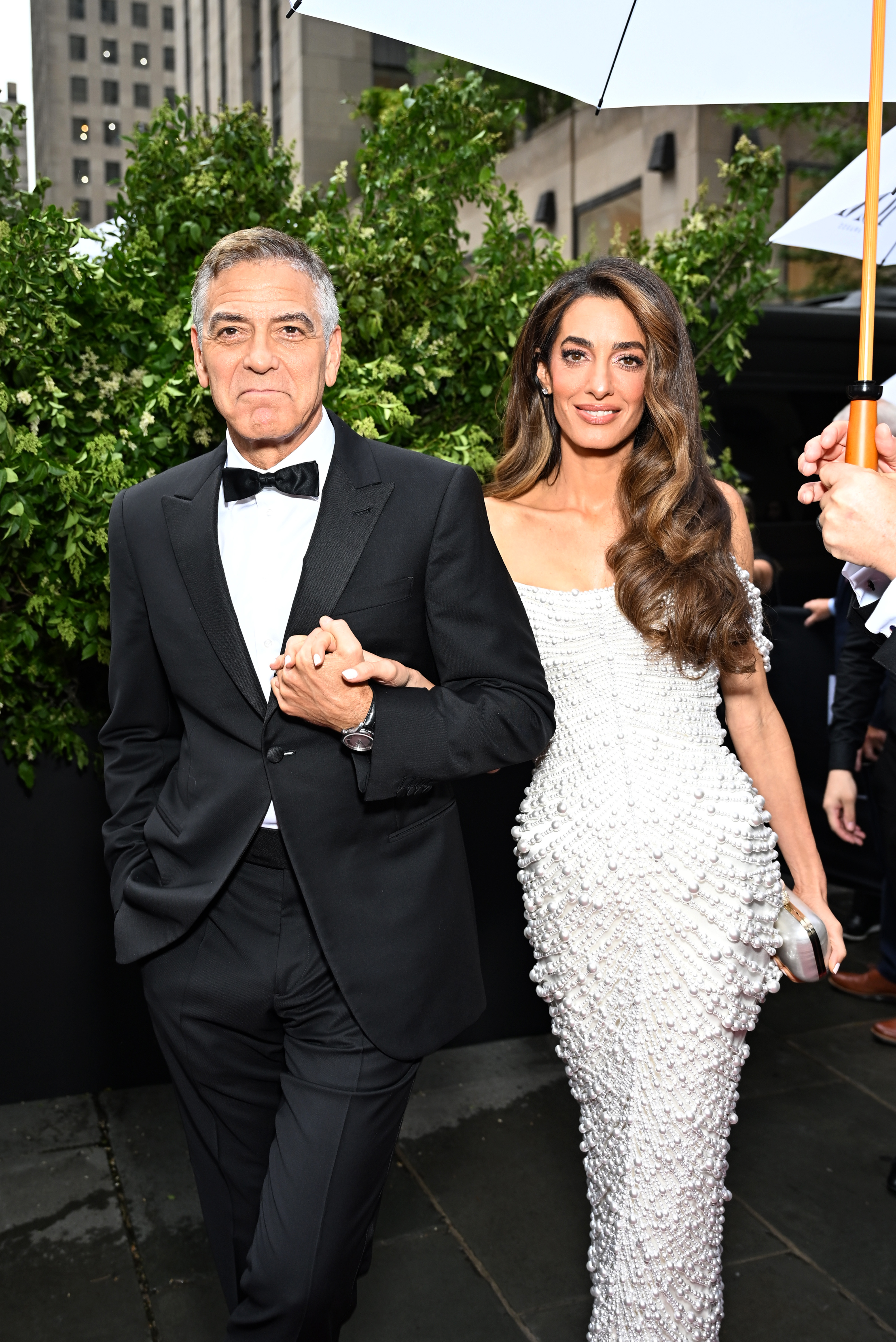 Walking arm in arm, George and Amal Clooney approach the entrance of the 78th Tony Awards, framed by greenery on a rainy day in Manhattan.