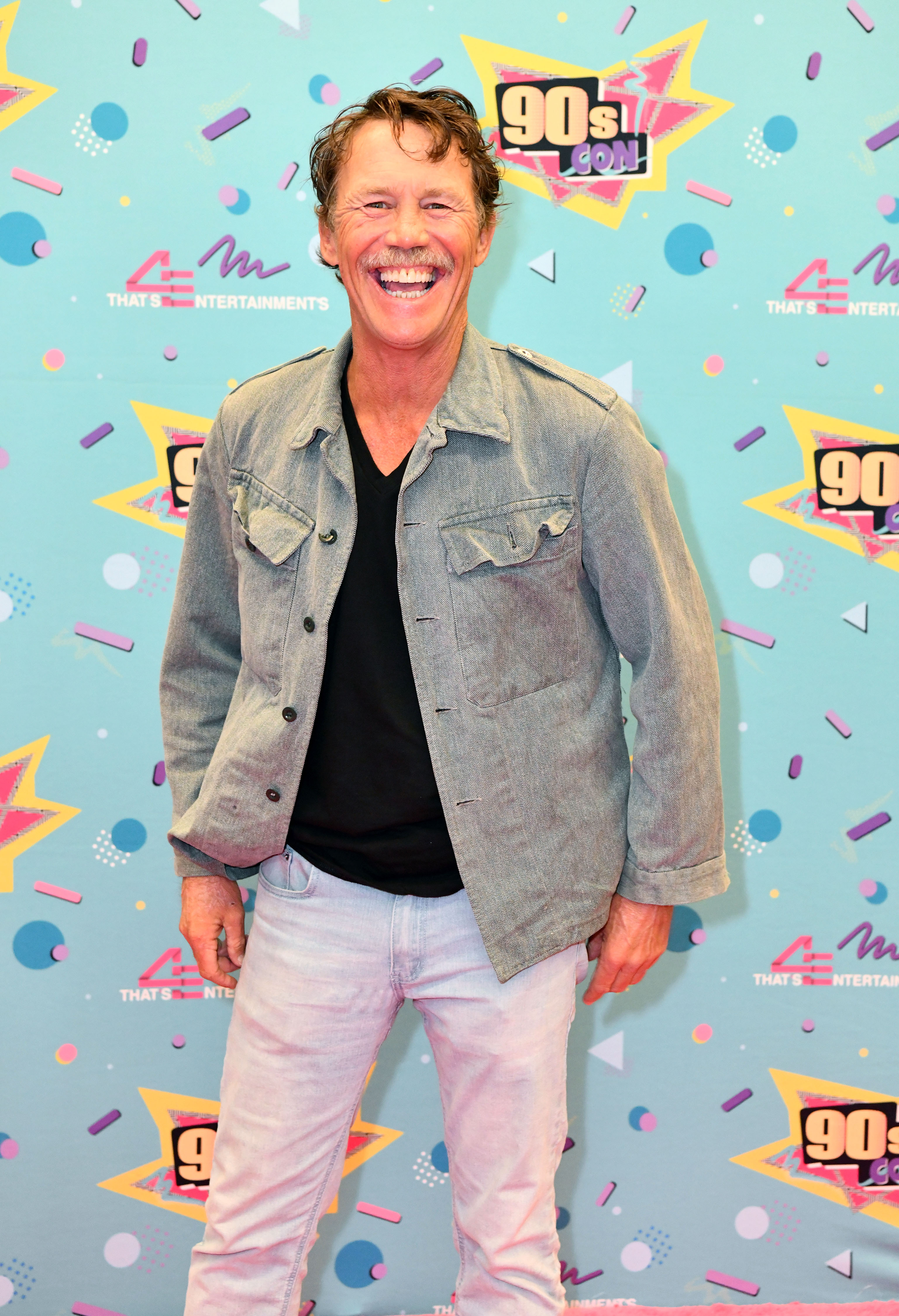Brian Krause attends 90s Con in 2024, posing on the red carpet with a wide smile. Dressed casually in a light denim jacket, black T-shirt, and pale jeans, he embraces the playful energy of the event. The colorful, retro-themed backdrop enhances the nostalgic tone of the appearance.