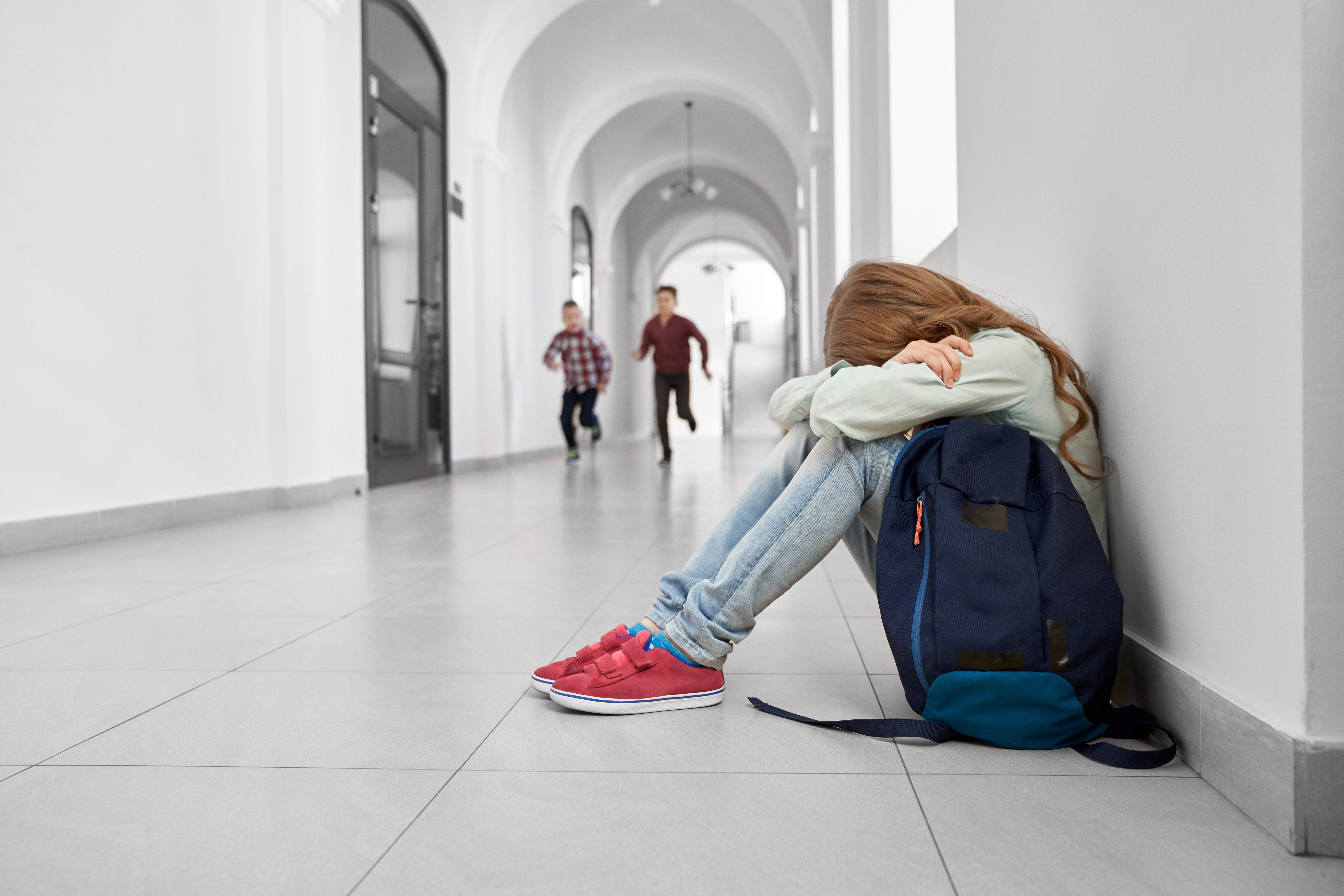 Mädchen sitzt in der Schule auf dem Boden und versteckt sich mit den Händen, als ihre Freunde im Hintergrund laufen und spielen. | Quelle: Shutterstock