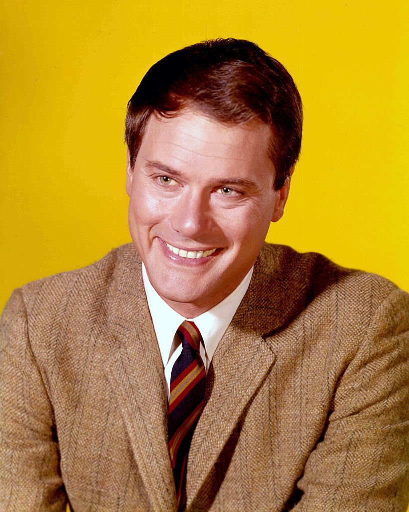 Larry Hagman, US actor, smiling, wearing a brown tweed jacket, a white shirt and a striped tie in a studio portrait, against a yellow background, issued as publicity for the US television series, 'I Dream of Jeannie', USA, circa 1967. | Source: Getty Images