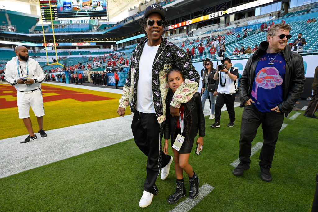 Jay-Z keeps a protective arm around Blue Ivy while crossing the field, surrounded by photographers and staff. The photo highlights a grounded family moment within the Super Bowl spectacle.