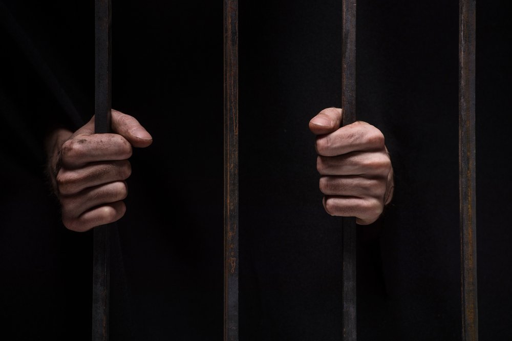 Gros plan sur les mains d'un homme derrière les barreaux de prison sur fond noir. | Shutterstock