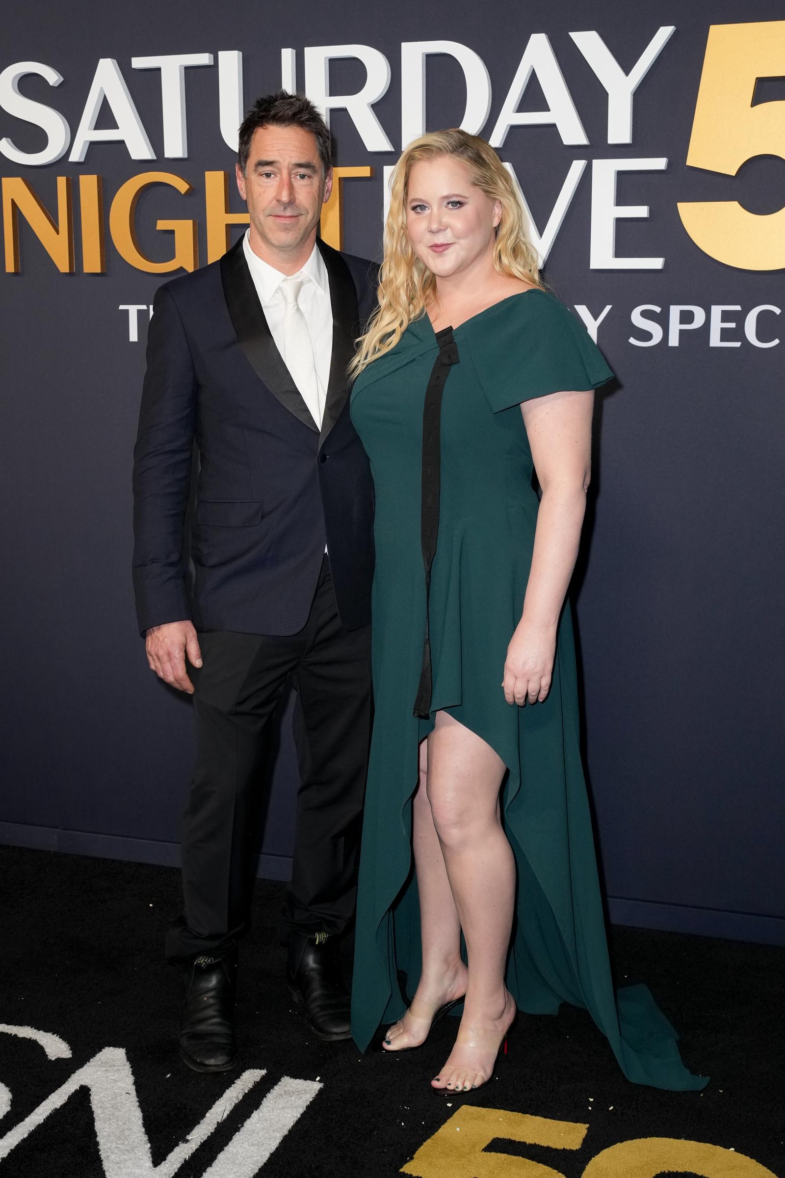 At SNL 50 on February 16, 2025, Amy Schumer wore a green gown with a cape sleeve and high slit as she posed with husband Chris Fischer, who wore a classic black tux with a white tie.