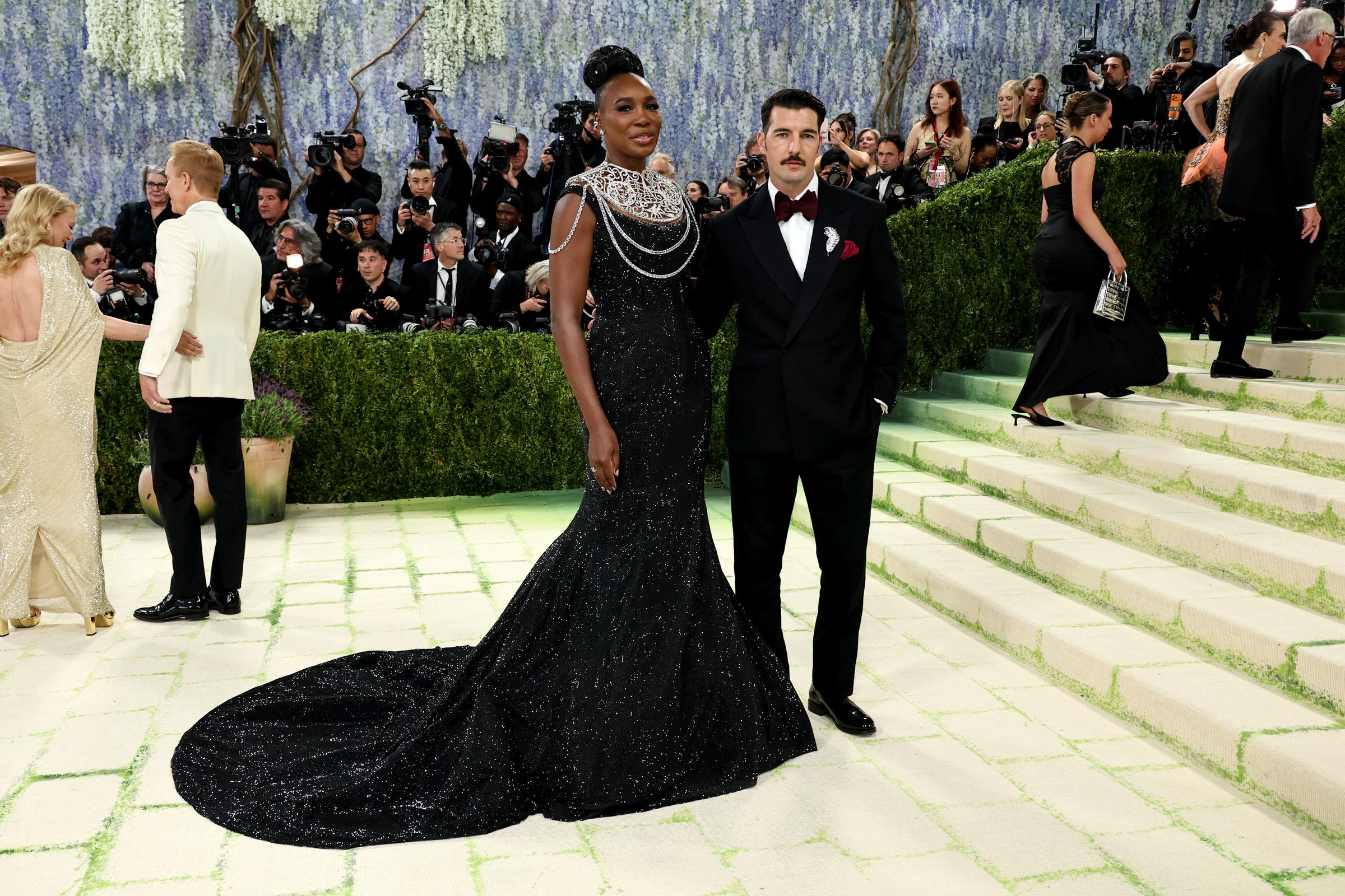 Venus Williams stuns in a fitted black gown with subtle sparkle and cascading pearl chains. The layered details add movement and a touch of elegance. Beside her, Andrea Preti keeps it sharp in a tailored black suit with a burgundy bow tie.