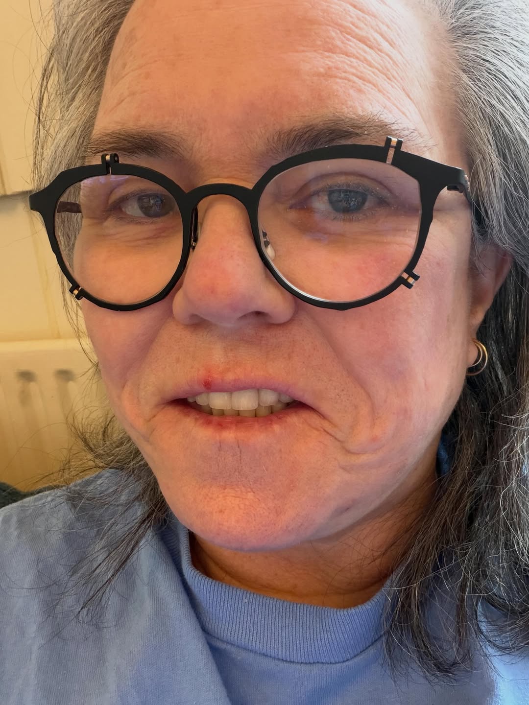 Rosie O'Donnell shares a close-up selfie while wearing black round glasses and a light blue top, posing in soft indoor lighting with a relaxed expression. | Source: Instagram/rosie