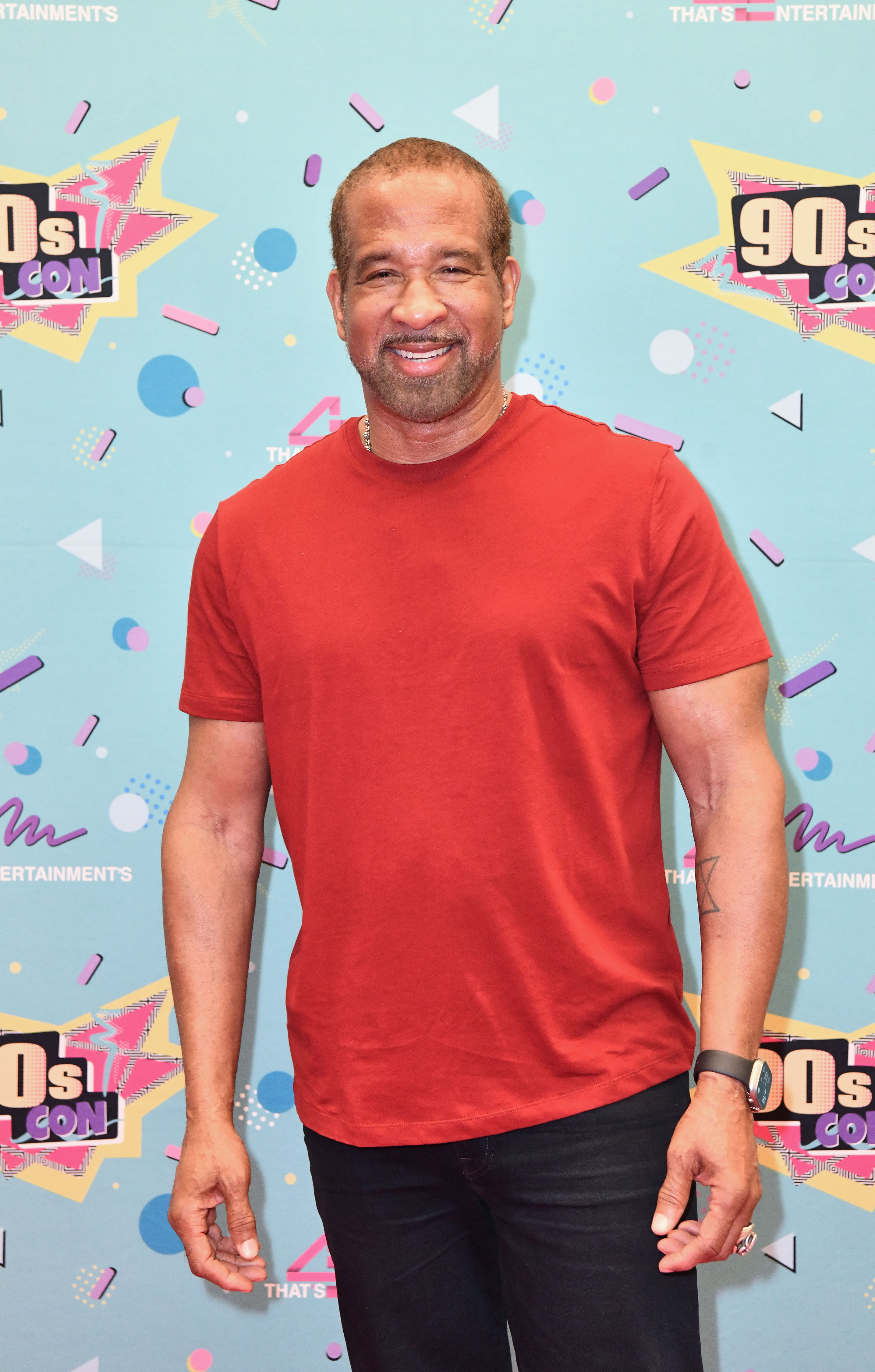 Dorian Gregory poses at 90s Con in 2024, wearing a casual red T-shirt and dark jeans. Smiling warmly in front of a colorful, retro-themed backdrop, he embraces the nostalgic energy of the event with a relaxed and approachable presence.