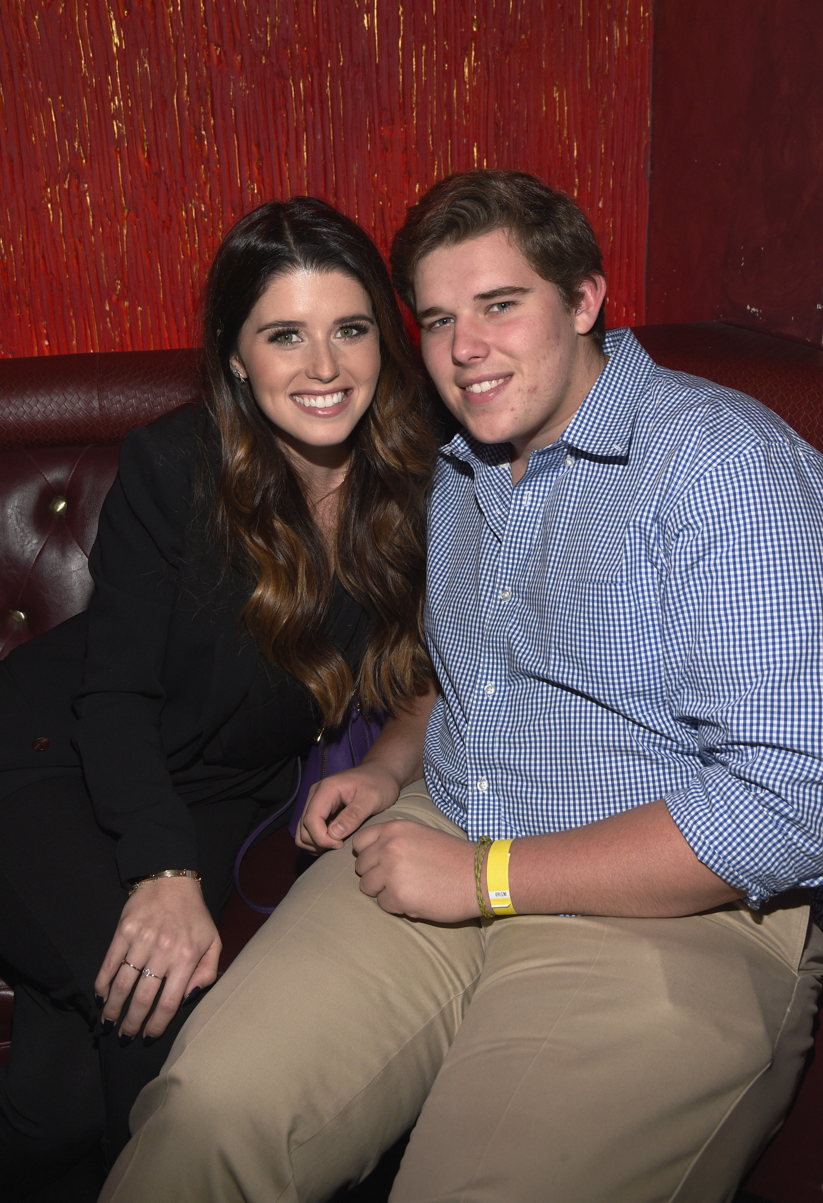 Christopher sits beside his sister Katherine at the pre-reception of an event in Los Angeles, in December 2014. He wears a blue checkered shirt and beige pants, with a yellow wristband visible on his right wrist. His rounder face and relaxed demeanor reflect his style during this period.