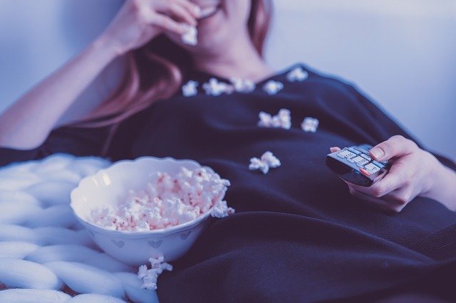 Woman sits on bed smiling and eating popcorn | Photo: Pixabay