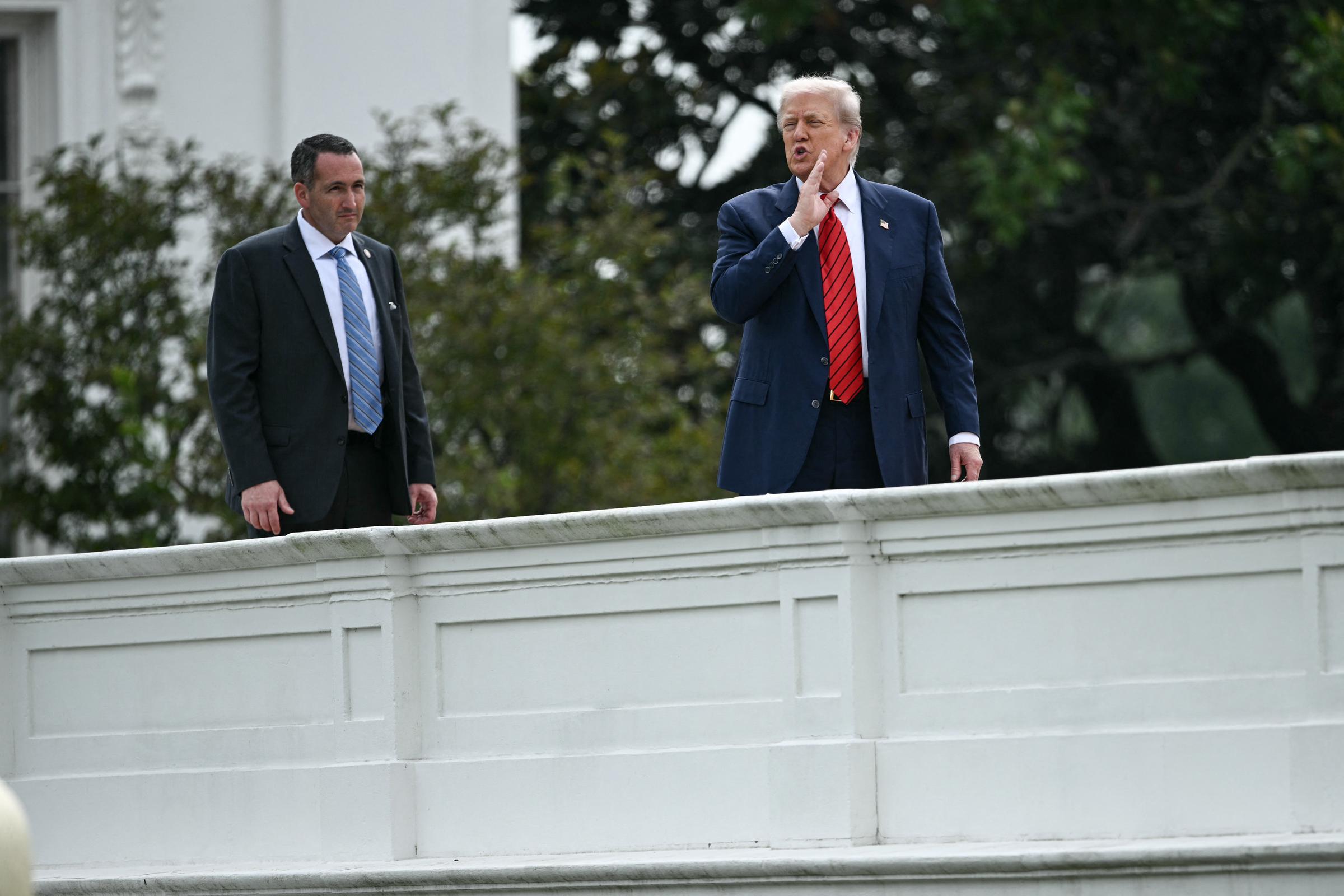 Donald Trump stands on the roof of the White House, one hand raised to his mouth as if calling out or addressing someone below, with a security detail member at his side.