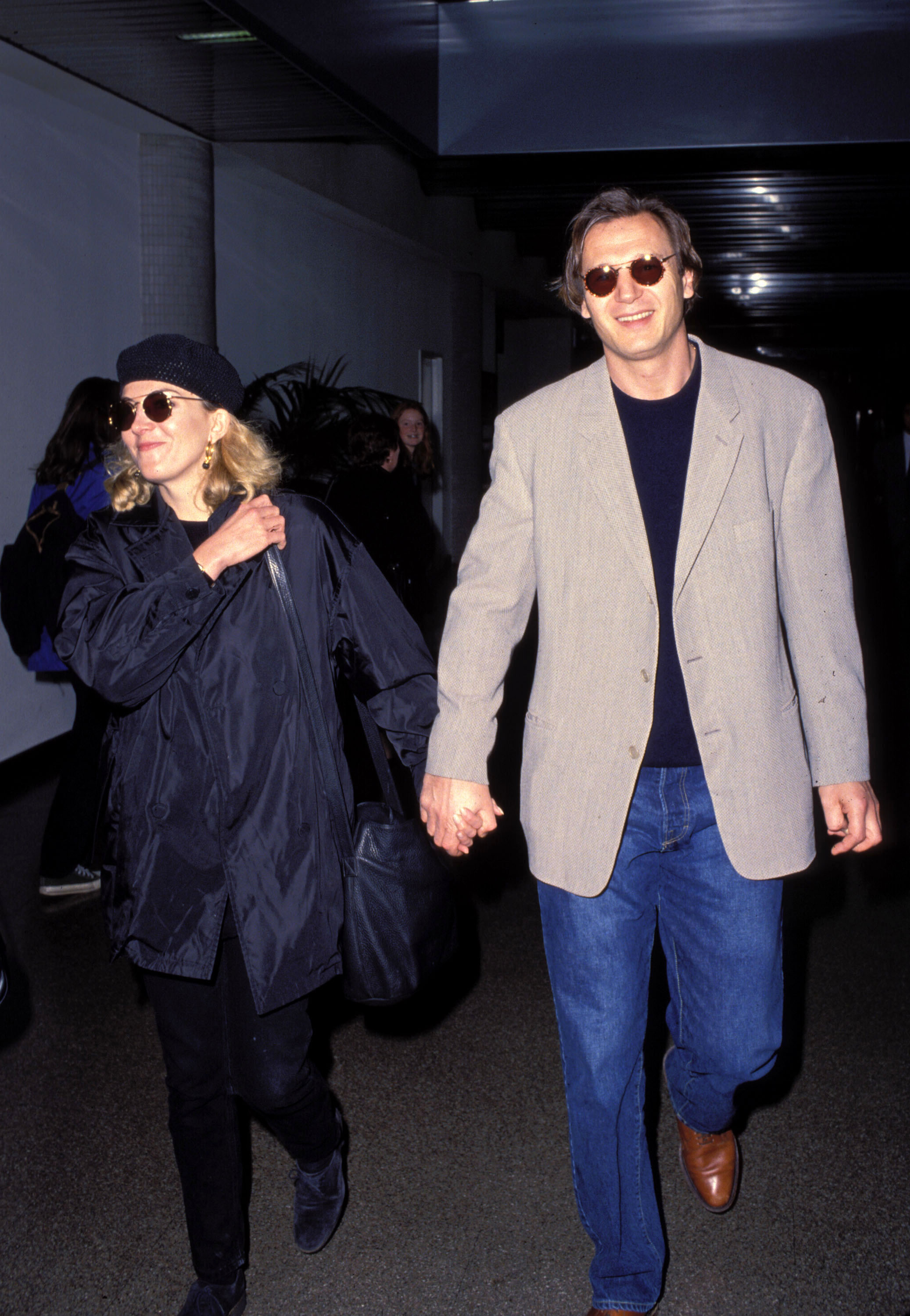 Natasha Richardson and Liam Neeson walk hand in hand through LAX Airport in Los Angeles on March 22, 1994, as they head to New York. Both wear sunglasses and appear relaxed while making their way through the terminal.