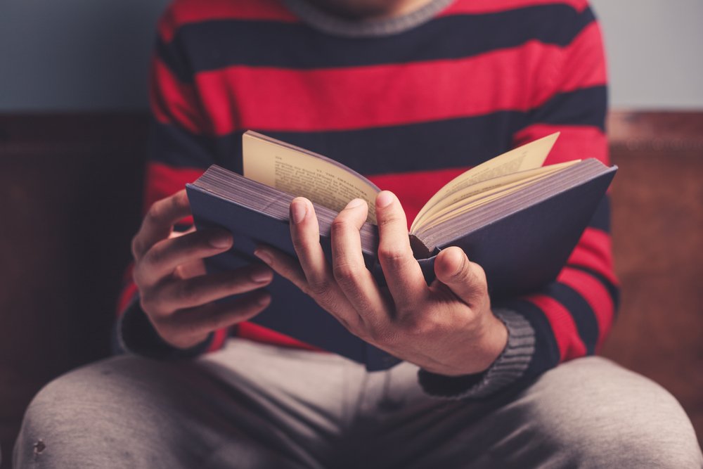 Joven sentado en un banquillo con un libro abierto entre sus manos. | Foto: Shutterstock