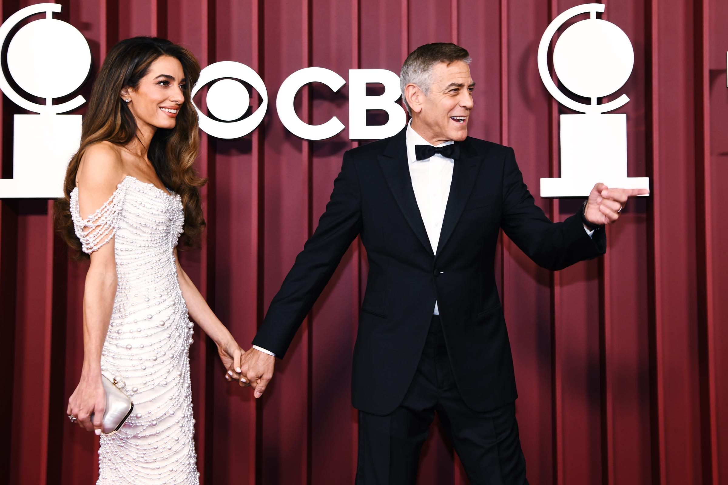 Amal Clooney glows as George Clooney chats expressively, their fingers interlaced on the red carpet. Her pearl-embellished gown dazzles beneath the lights, while he keeps the mood lively with a playful gesture.