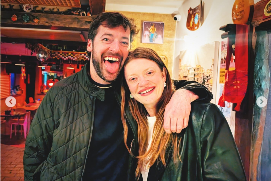Two guests stand close together inside a warmly lit space at Jack Quaid and Claudia Doumit’s wedding, smiling broadly as the man playfully sticks out his tongue. | Source: Instagram/smokey_horse