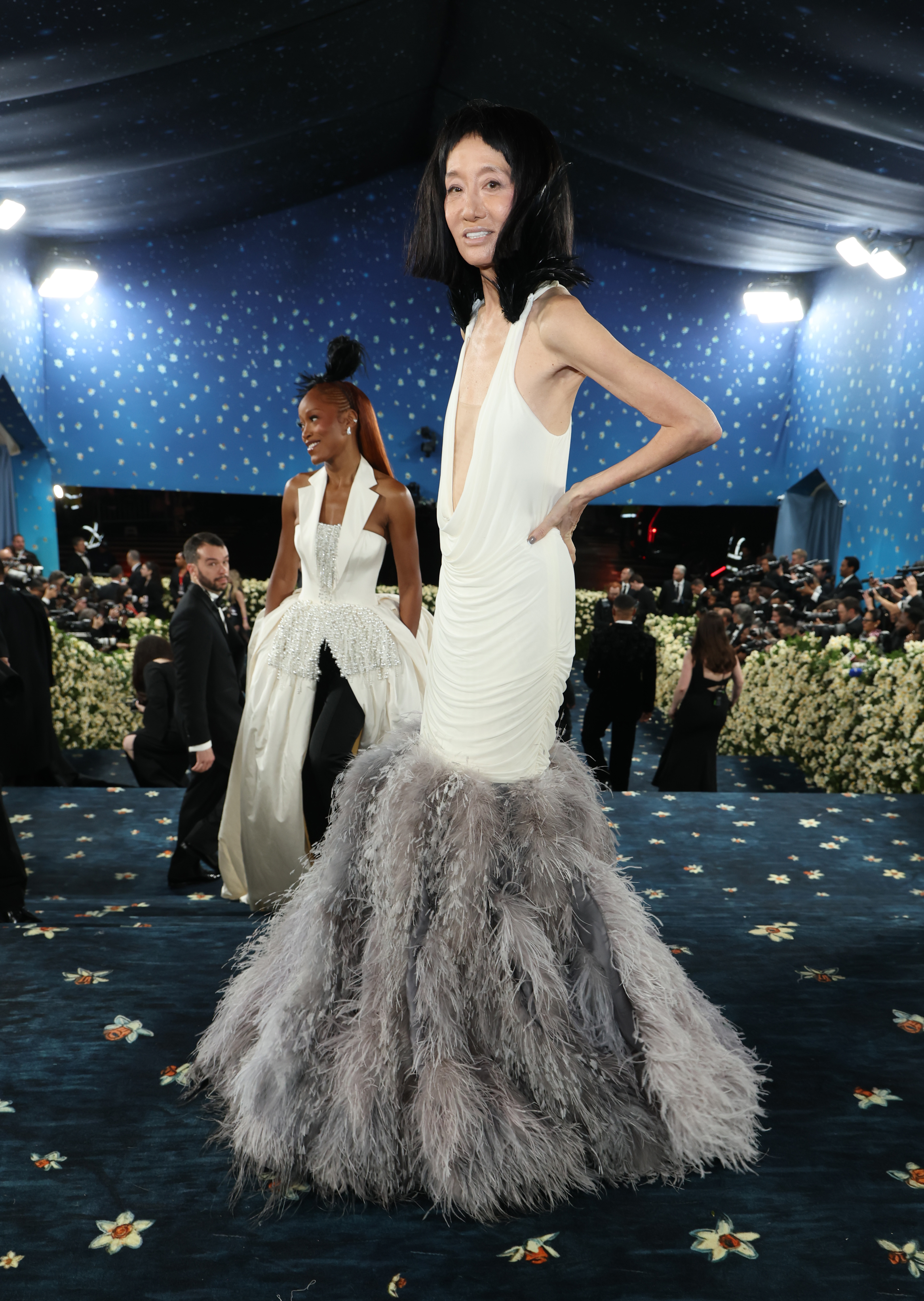 Surrounded by a sea of cameras and floral decor, Vera Wang poses mid-step in her textured gown as Keke Palmer follows behind, both dressed in striking white looks with feathered flair.