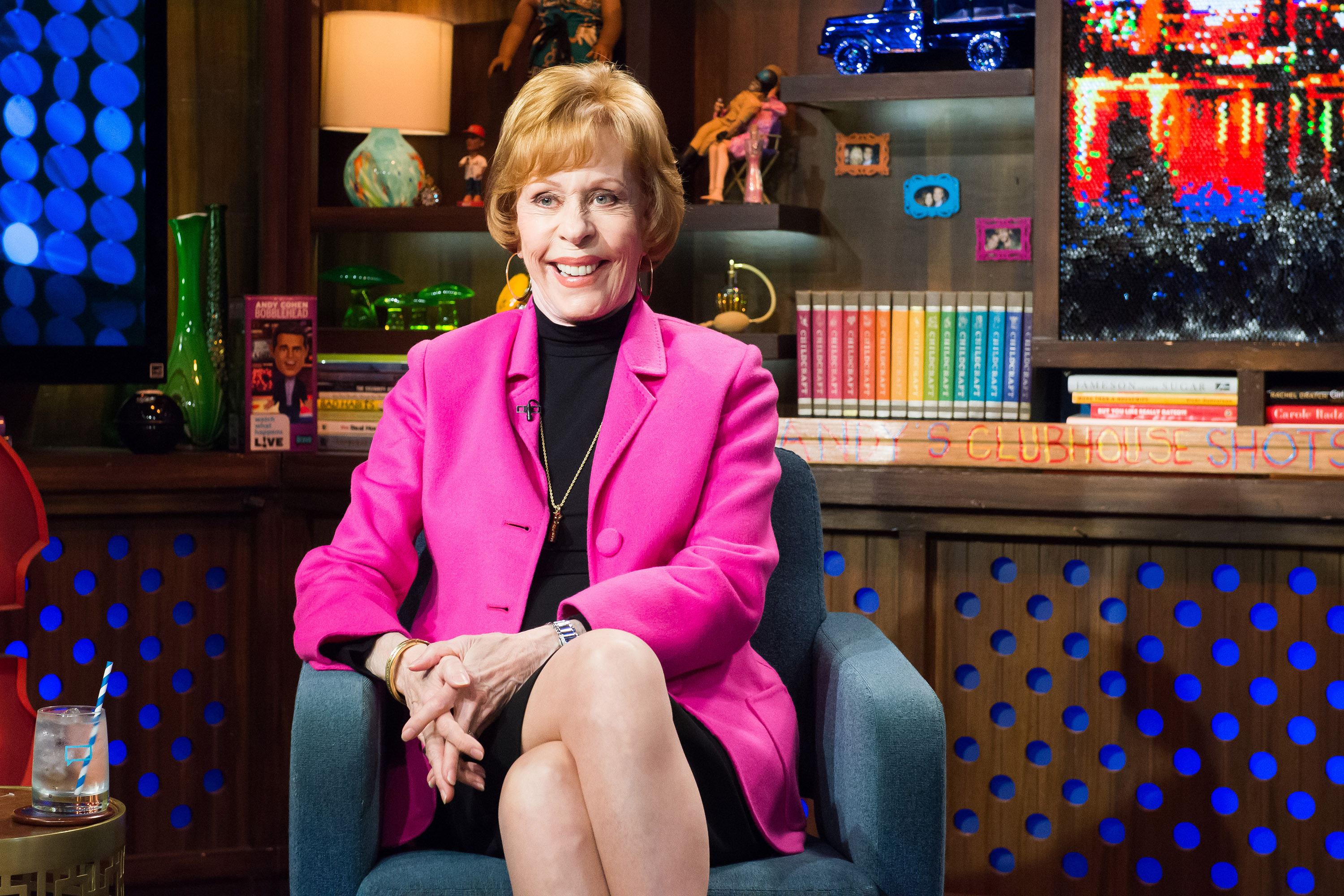 Seated in a studio chair, she wears a bright pink blazer over a black turtleneck dress. Her legs are crossed, and her hands rest neatly on her knee. The set behind her features shelves filled with books, decorative objects, and colorful lighting, giving the space a lively, layered look.