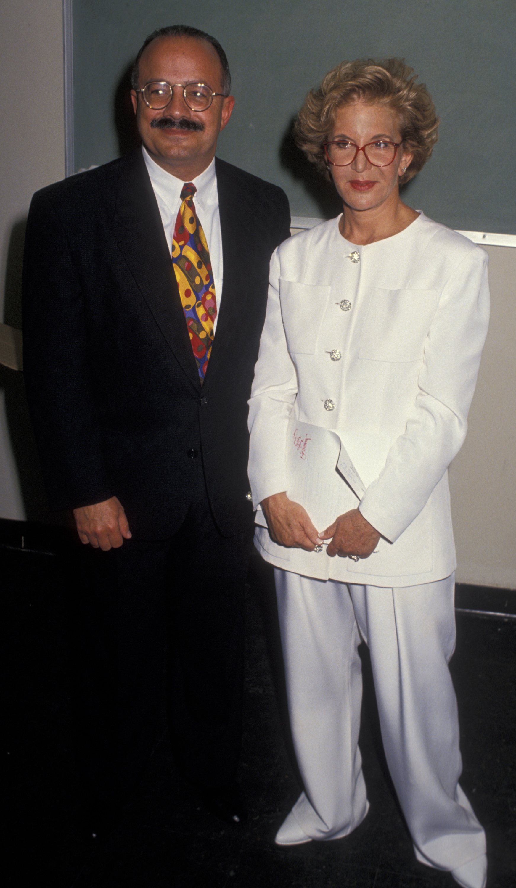 Sally Jessy Raphael and her husband, Karl Soderlund, attend the Art Against AIDS IV Benefit Auction at the Biltmore Hotel in Coral Gables, Florida, on April 10, 1994. Raphael wears a crisp white pantsuit with crystal buttons and her iconic red glasses, while Soderlund stands beside her in a colorful tie and dark suit.