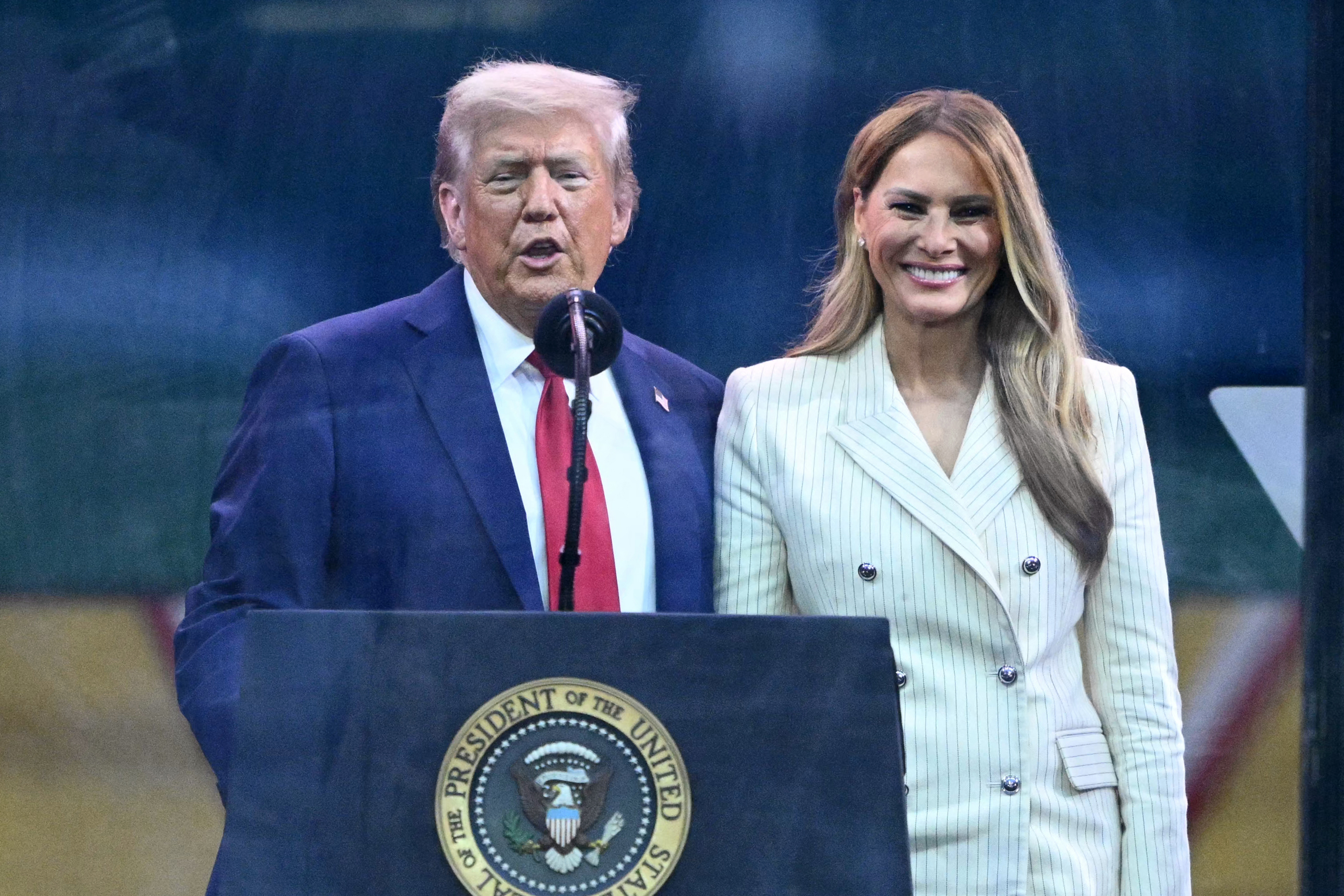 Donald Trump delivers remarks beside Melania Trump following the US Army’s 250th anniversary parade. Standing behind the presidential seal, he speaks into the microphone while Melania smiles beside him.