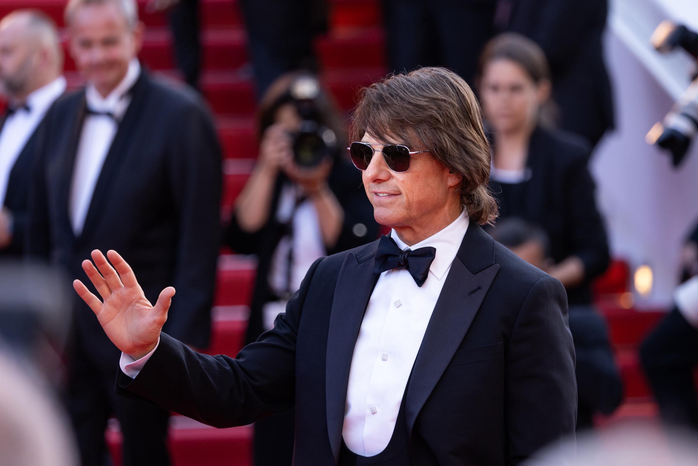 Tom Cruise flashes a relaxed smile and waves to the press as he makes his entrance at the "Mission: Impossible – The Final Reckoning" red carpet premiere in Cannes. His formal black tuxedo look was paired with tinted sunglasses and his signature charm.