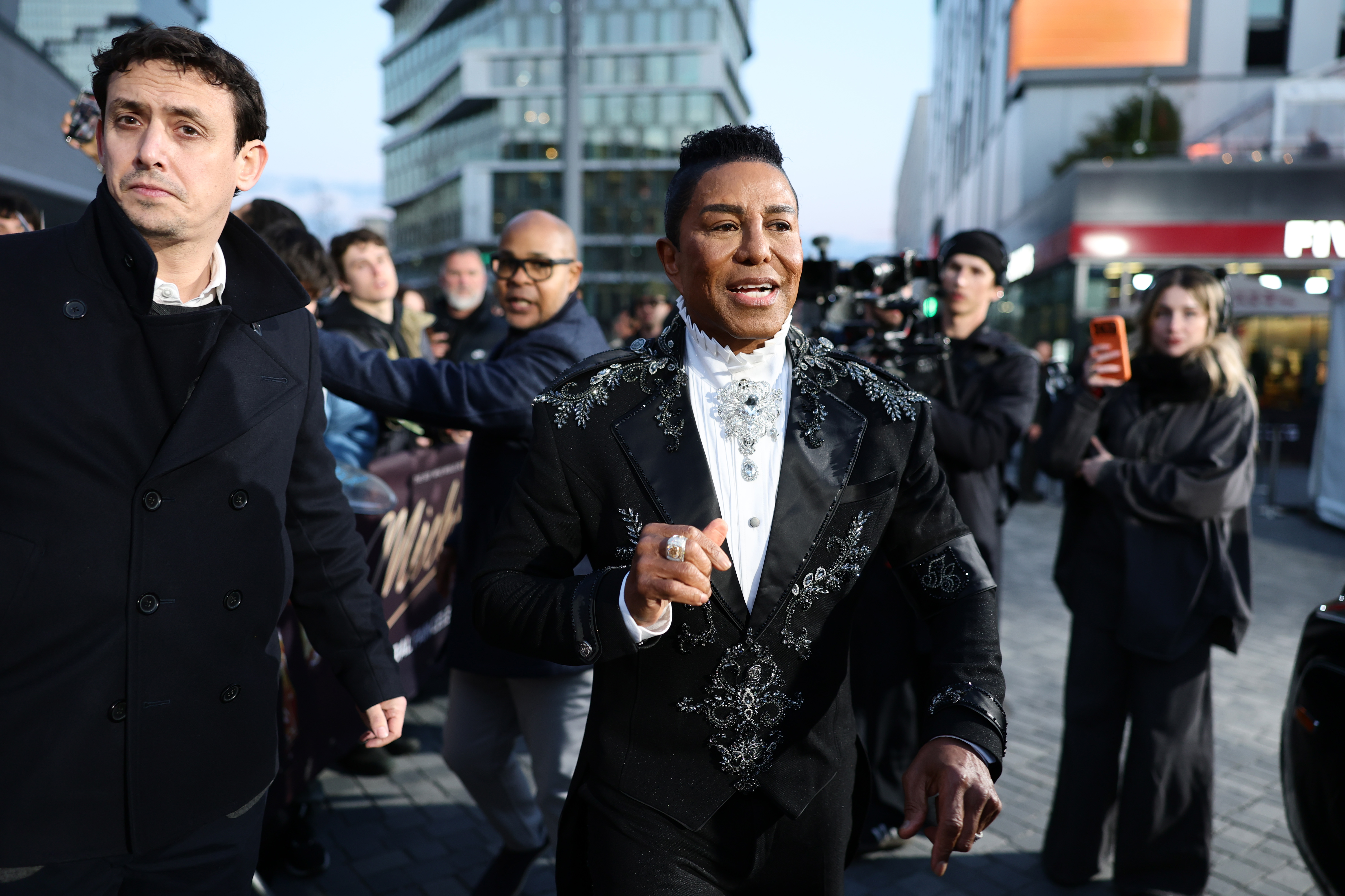 Jermaine Jackson arrives at the "Michael" Global Fan Premiere in Berlin, commanding attention in a sharply tailored black suit adorned with intricate silver embellishments. Moving through the crowd with cameras flashing, his regal styling and confident stride give the moment a grand, almost theatrical presence.