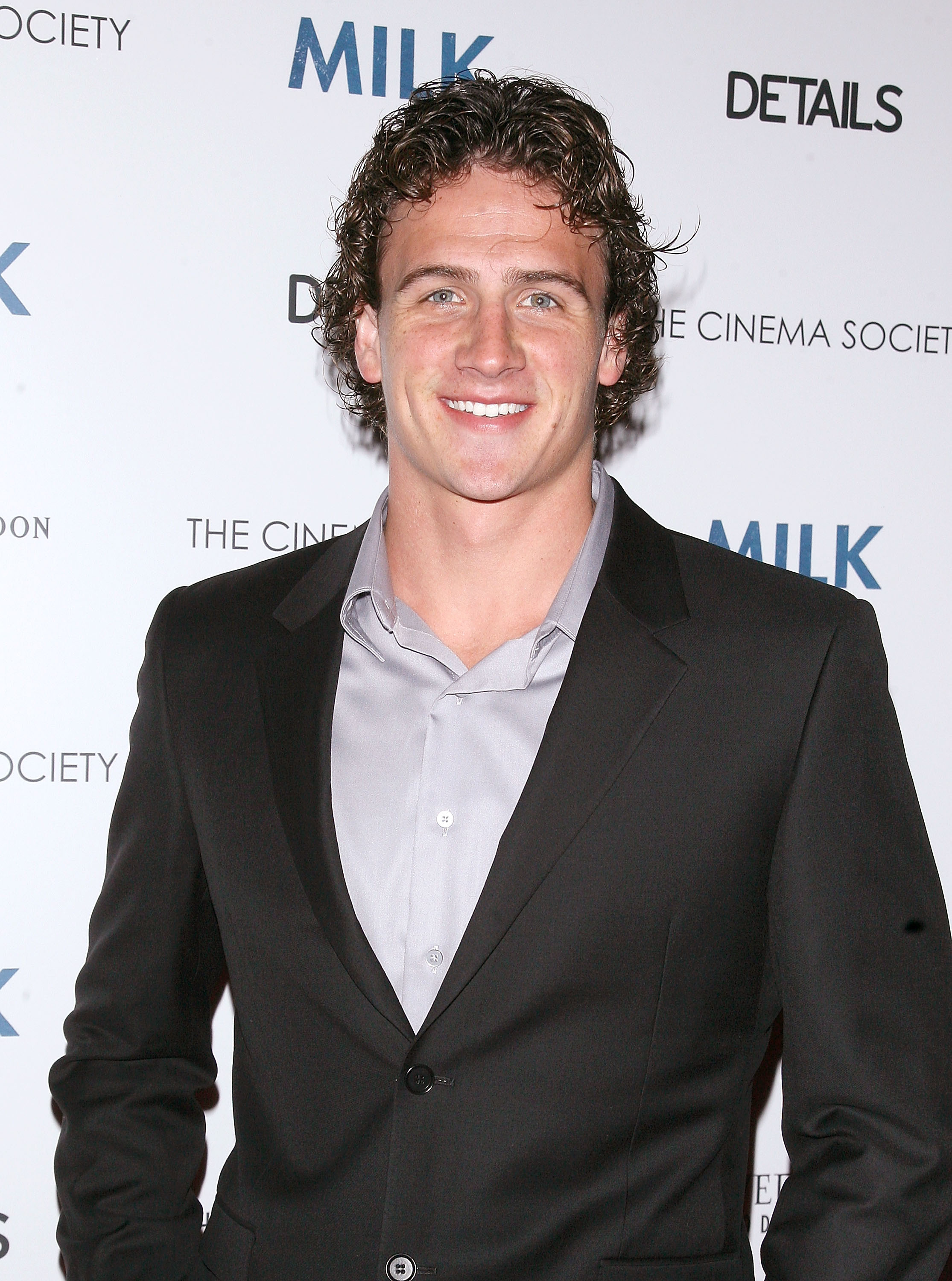 Ryan Lochte smiles for photographers while attending a New York City screening of "Milk," dressed in a dark tailored suit over a light gray shirt. The Olympic swimmer poses confidently against a step-and-repeat backdrop at the Landmark Sunshine Theater.