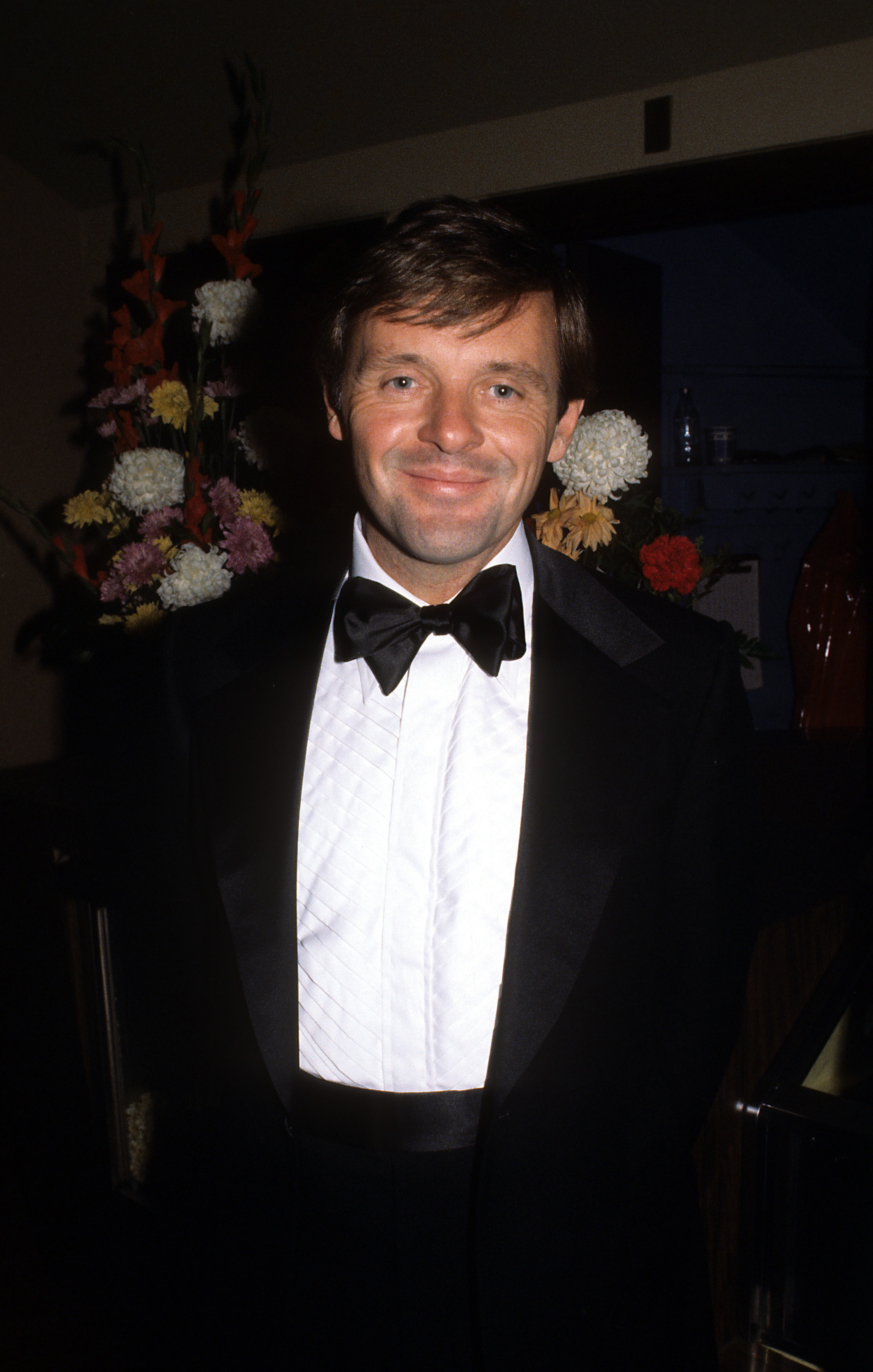 Anthony Hopkins beams warmly in an elegant black tuxedo, his charm illuminated by soft evening light. The neatly tied bow and crisp white shirt add a touch of classic poise. Surrounded by vibrant flowers, he radiates confidence and timeless grace.