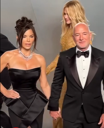 Lauren Sánchez stands with one hand on her hip while holding hands with Jeff Bezos on the red carpet as Nicole Kidman walks past behind them in a shimmering gold gown. | Source: Facebook/NewYorkPost