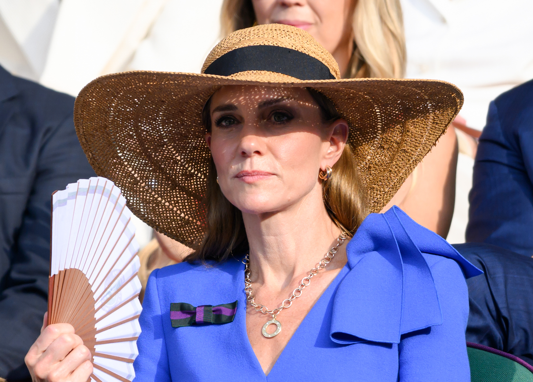 Princess Catherine holds a fan in one hand as she watches intently from the Royal Box. The brim of her hat casts a patterned shadow across her face in the afternoon light.