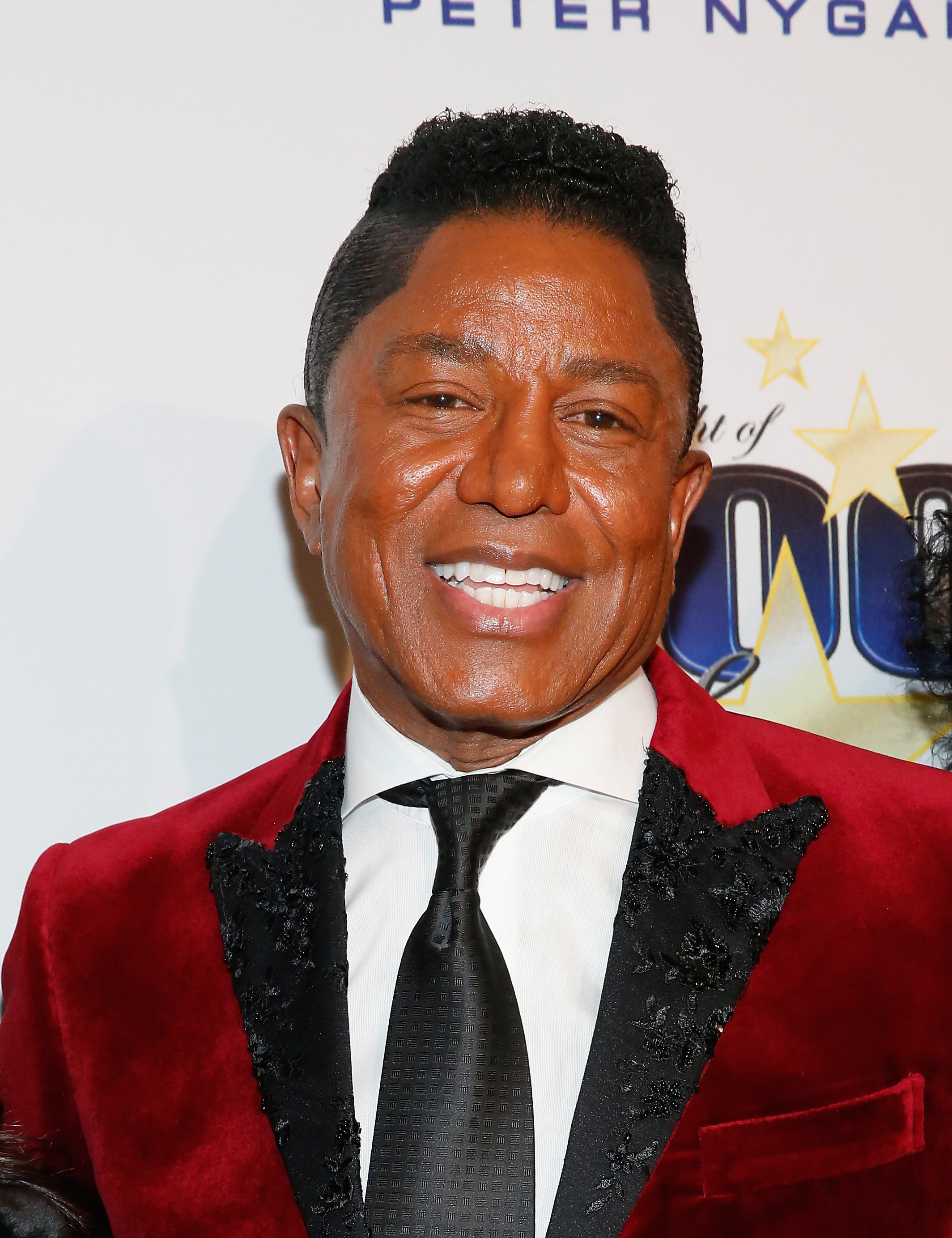 Jermaine Jackson smiles brightly at the Night of 100 Stars Oscar Viewing Gala, wearing a rich red velvet jacket with black detailing. The bold texture and classic tie pairing create a standout red carpet look that blends sophistication with flair.