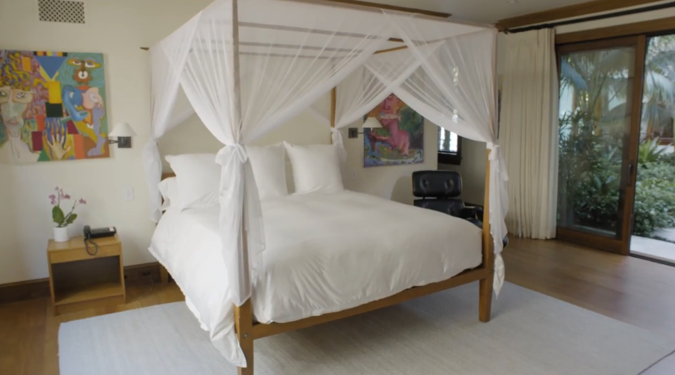 Sheer white netting drapes gracefully over a wooden four-poster bed, contrasting bright, colorful artwork hanging on the crisp white walls. Sliding glass doors beside the bed open directly out to a garden patio surrounded by lush tropical greenery. | Source: YouTube/Architectural Digest