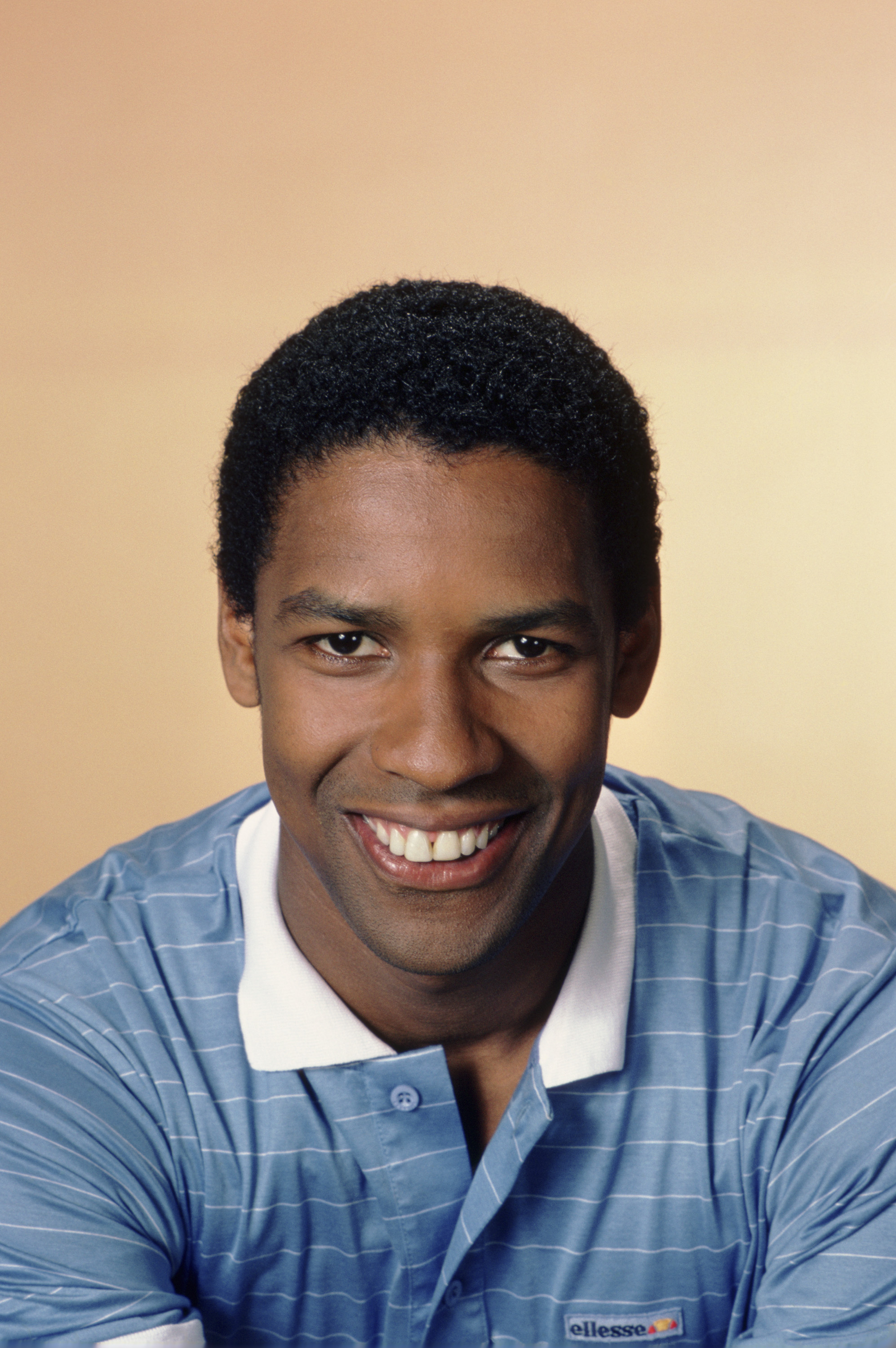 Denzel Washington poses in a powder blue polo during his early career. Sporting a low/small afro and clean-shaven look, this portrait captures the rising star's charm while filming "St. Elsewhere."