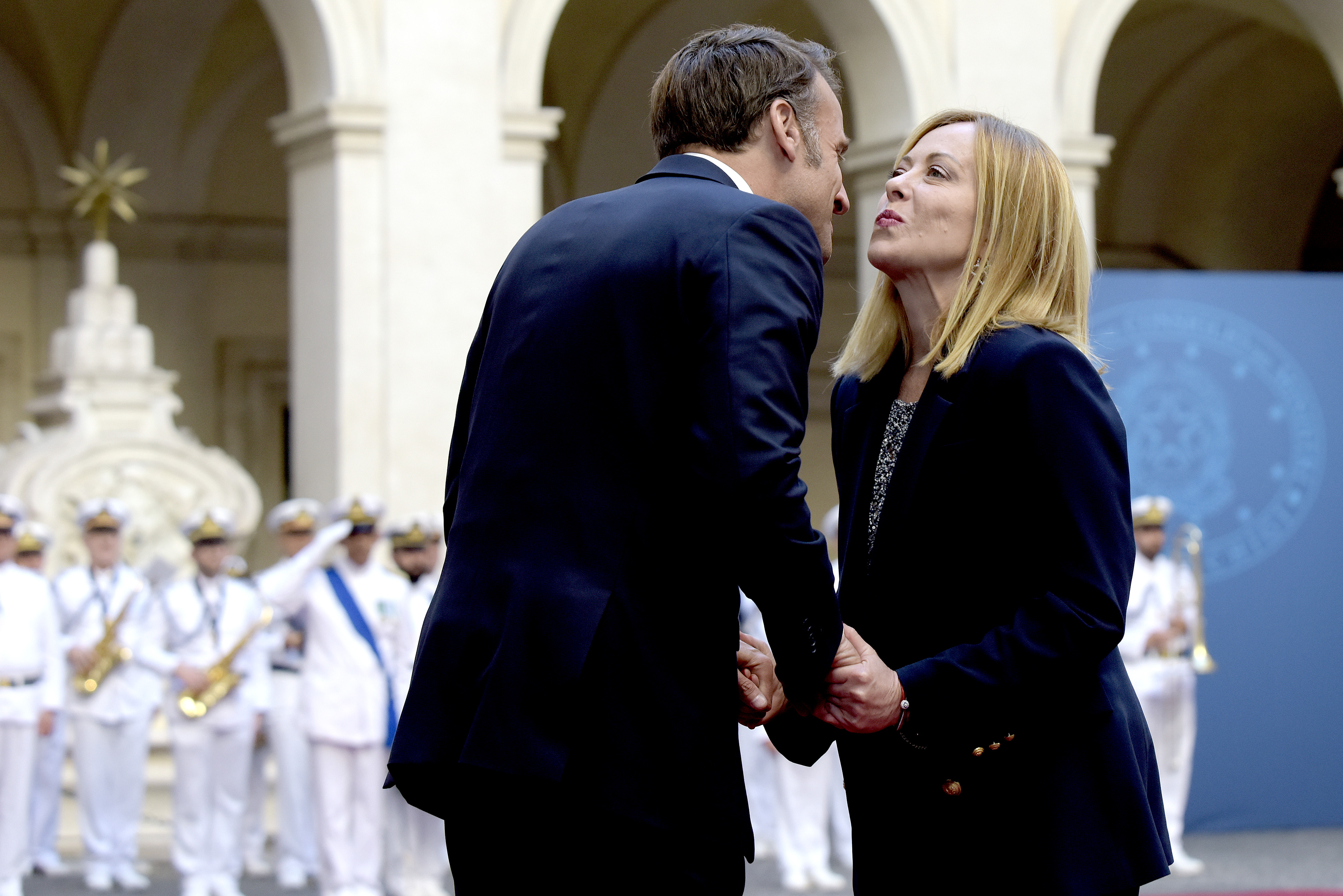 Emmanuel Macron and Giorgia Meloni lean in closely as they exchange greetings at Palazzo Chigi, holding hands as they stand face to face in front of a line of uniformed officials.