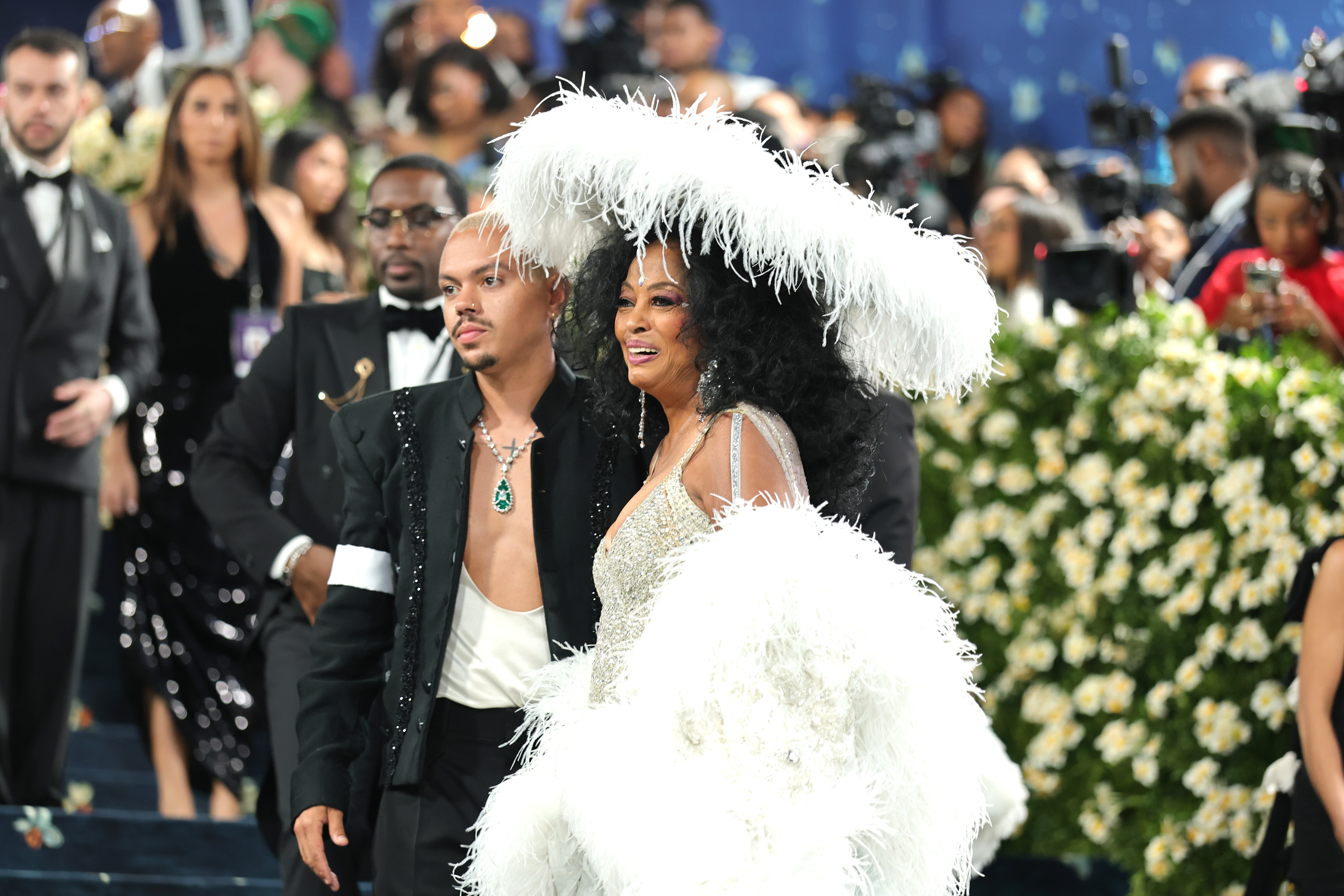 In a closer shot, Evan Ross’s open neckline reveals a cross tattoo on his chest and a necklace with a striking emerald drop resting against his neck. Beside him, his mother smiles sweetly, her expression warm and proud.