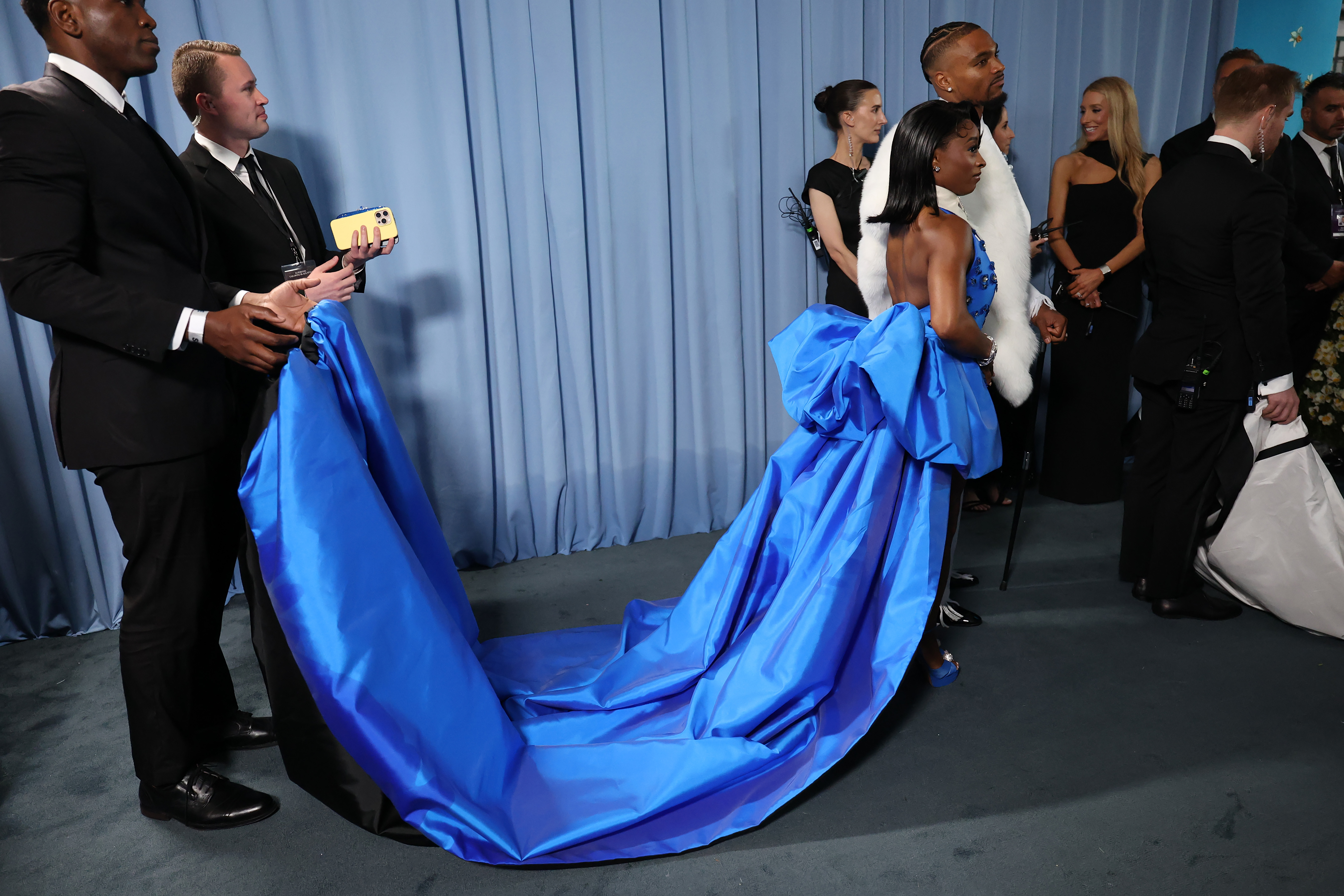 A staff member assists Simone Biles with her voluminous train as she walks forward holding Jonathan Owens' hand. Their posture remains calm amid the bustle.