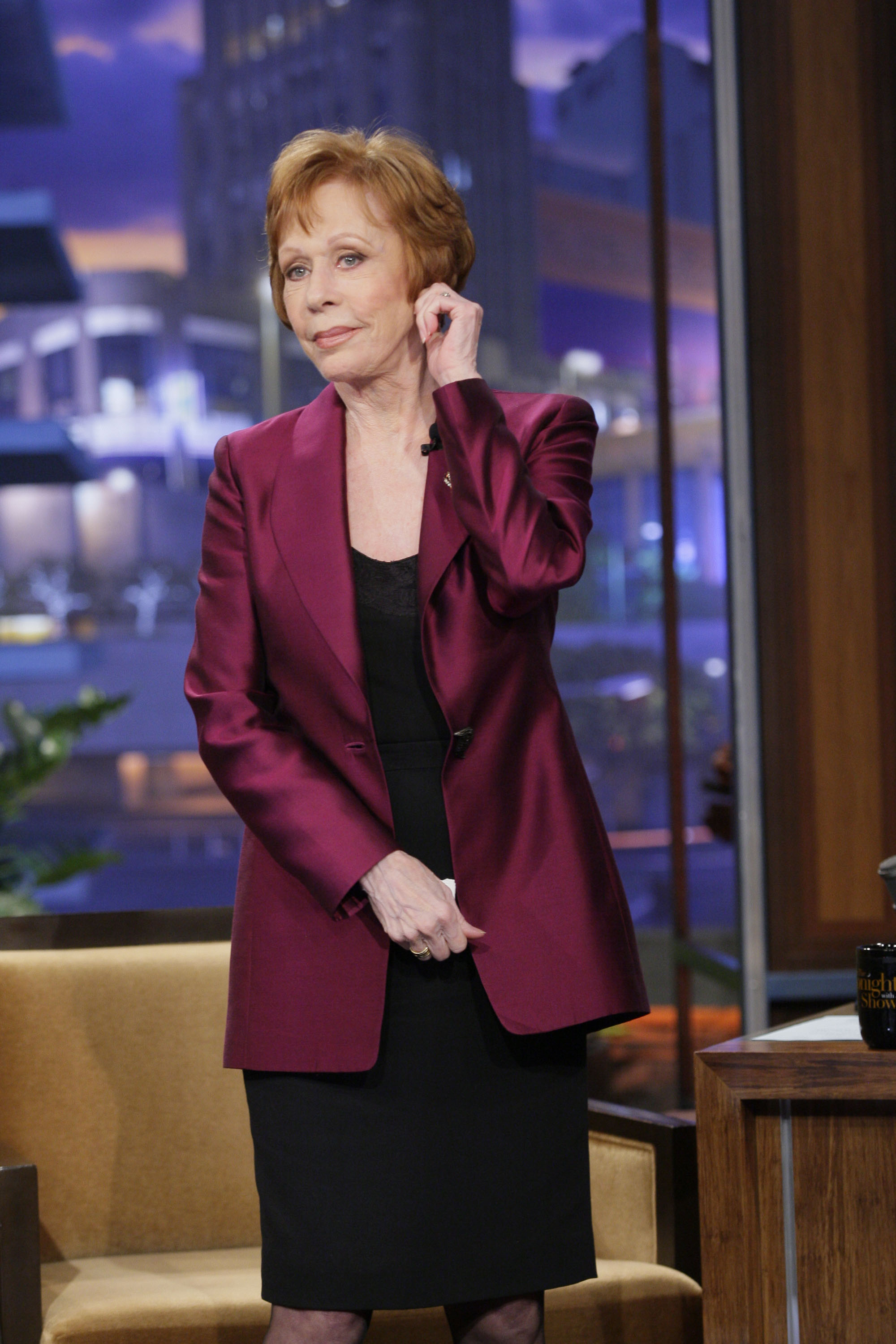 During a talk show appearance, she stands beside a chair wearing a deep burgundy blazer over a black dress. One hand reaches up toward her ear while the other rests near her waist, with a studio set behind her that includes a desk and cityscape backdrop visible through large windows.
