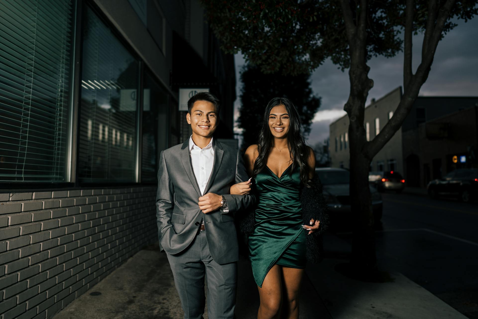 Una joven pareja feliz, elegantemente vestida, pasea del brazo por una calle de la ciudad por la noche | Fuente: Pexels