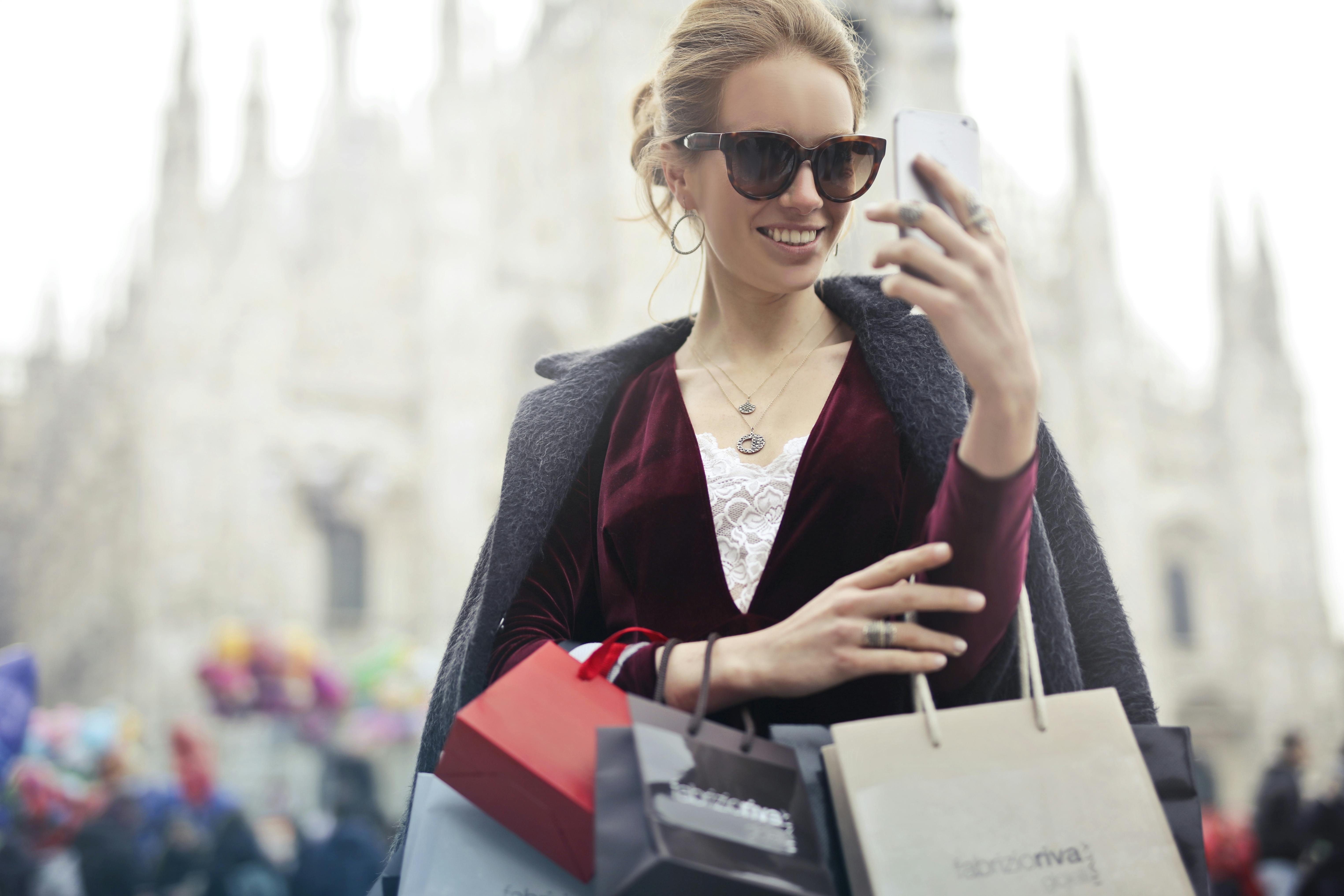 A wealthy woman wearing sunglasses, holding shopping bags, and looking at her phone | Source: Pexels