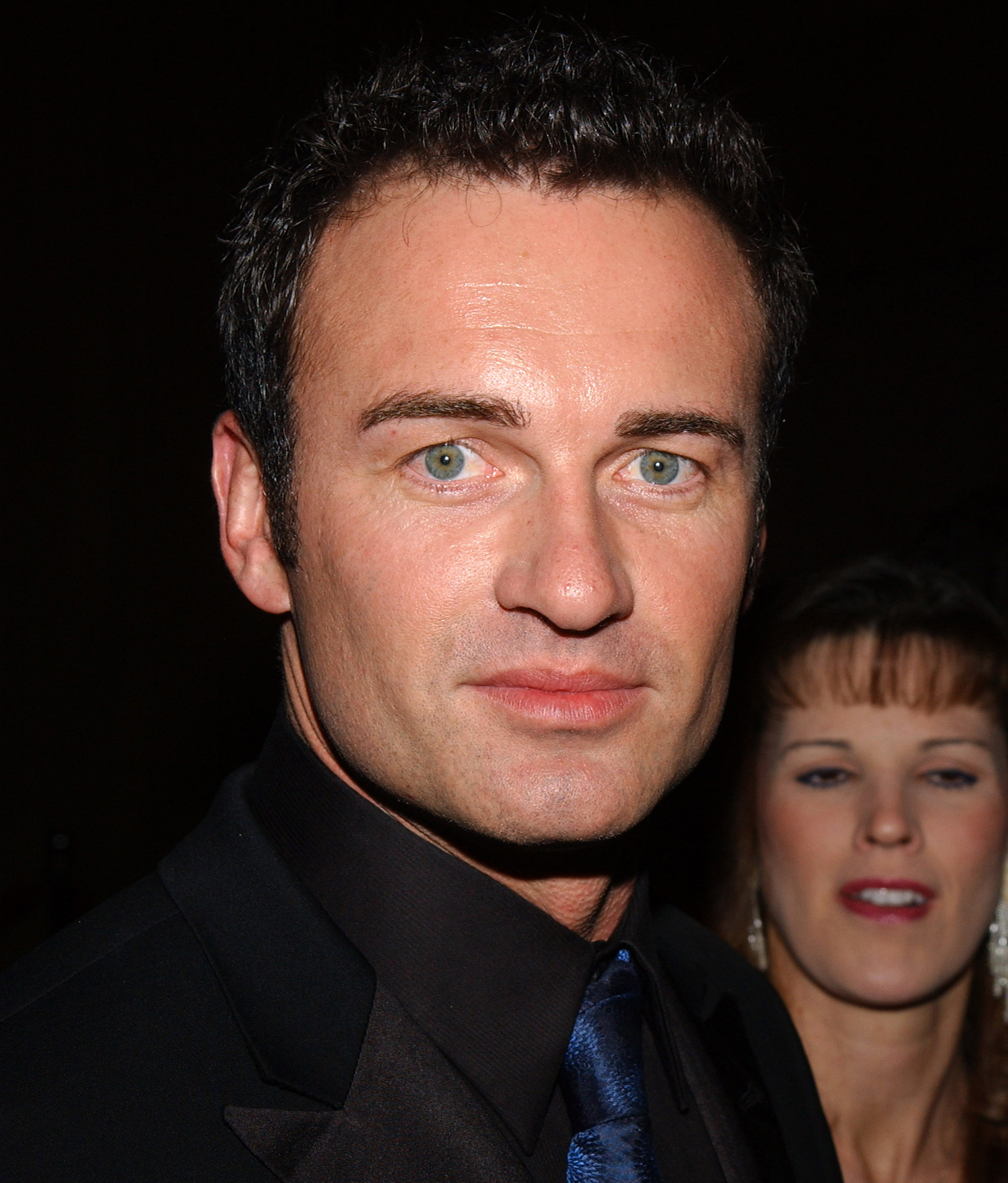 Julian McMahon is seen at a formal event in the early 2000s, wearing a black suit and dark shirt paired with a deep blue satin tie. His direct gaze and confident expression are sharply captured in the foreground, while the softly blurred background highlights the evening's social setting.