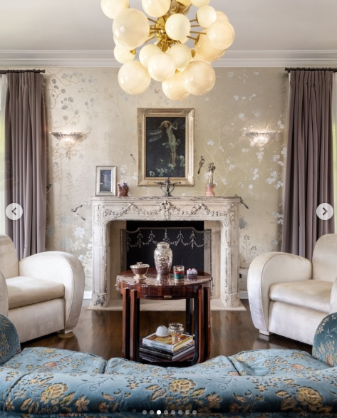 A sitting room with hand-painted floral wallcoverings frames an ornate carved marble fireplace, topped with a gilded oil painting of a reclining figure. Paired cream armchairs flank a tiered walnut side table, whilst a teal brocade sofa anchors the foreground and a cluster globe chandelier casts a warm, diffused glow across the room. | Source: Instagram/top.music.songs
