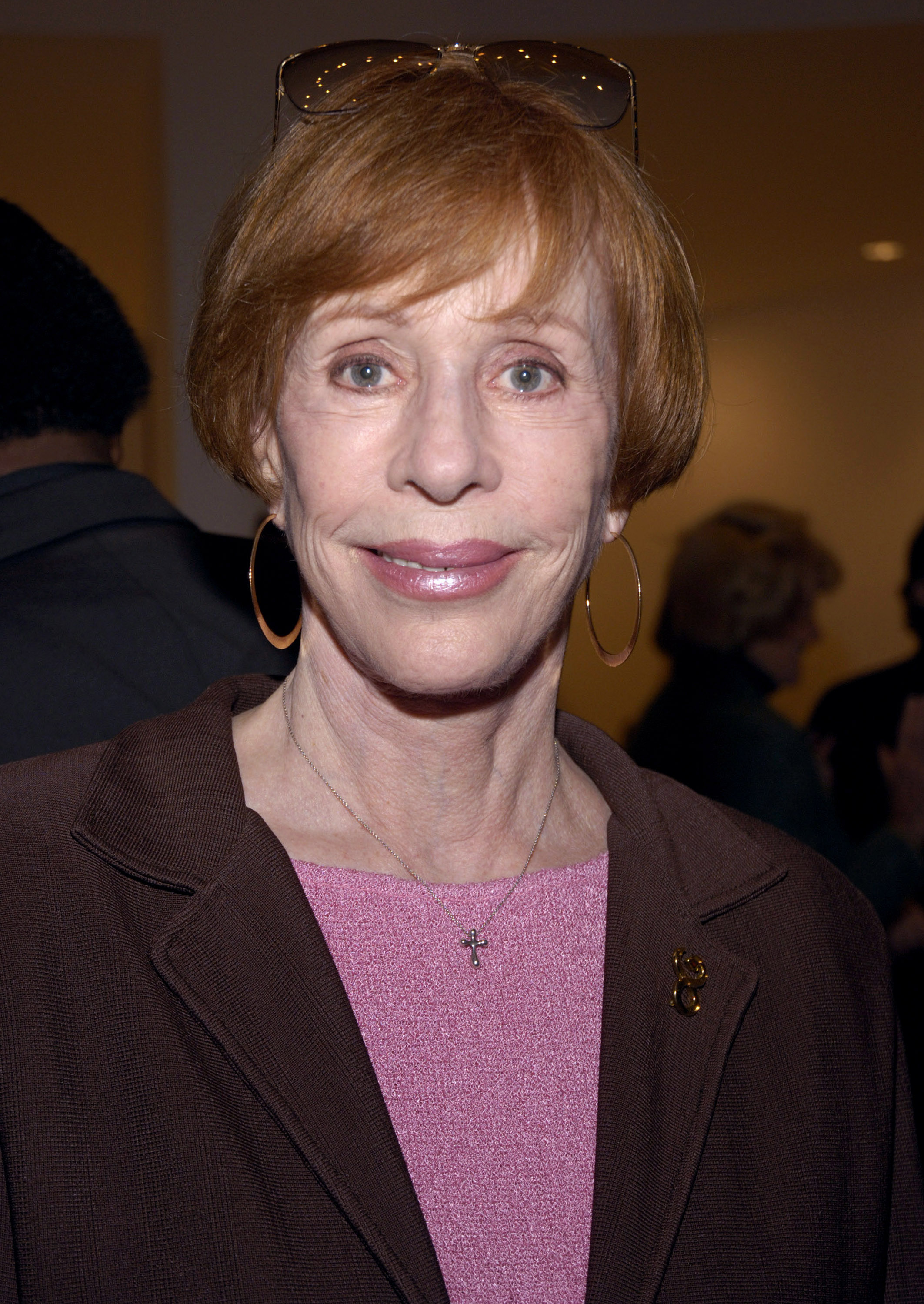 She appears in a close-up wearing a soft pink knit top under a brown blazer. Gold hoop earrings and a delicate necklace with a small cross pendant complete the look, while sunglasses rest on top of her short, light-brown hair.