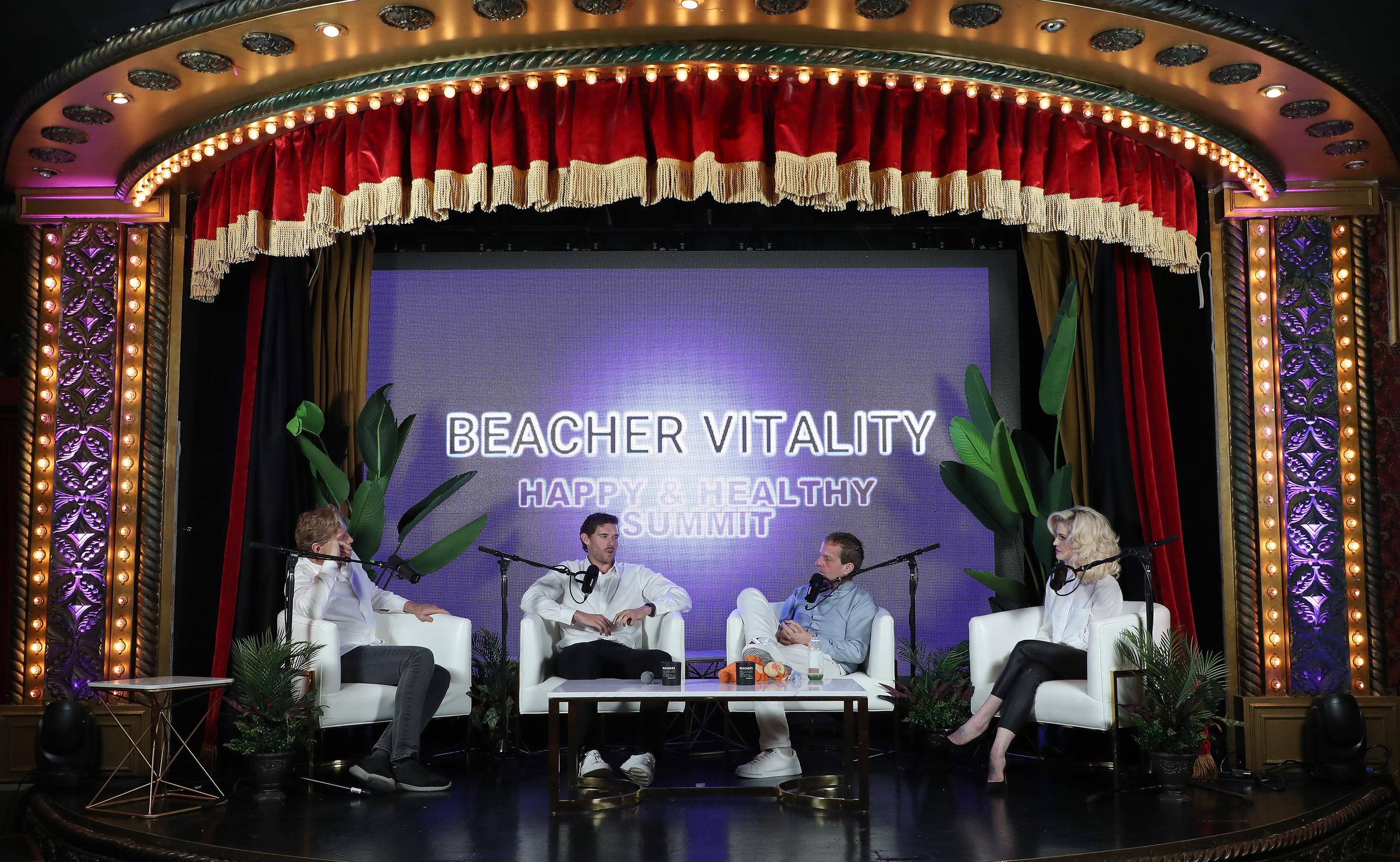 Under the ornate lights of The Hollywood Roosevelt stage, Christopher Schwarzenegger joins fellow speakers for a discussion centered on health and well-being. The panel setting features vibrant lighting and tropical décor, setting the tone for the day's uplifting theme.