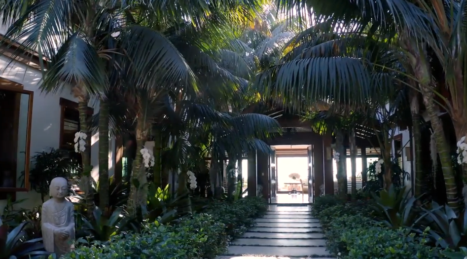 Lush palm trees create a dense, verdant canopy over the garden courtyard, shading a stone-paved walkway that leads directly toward the residence’s open entryway. White orchids mounted on palm trunks add subtle floral details to the scenery, while a calm stone Buddha statue sits near the garden bed on the left. | Source: YouTube/Architectural Digest
