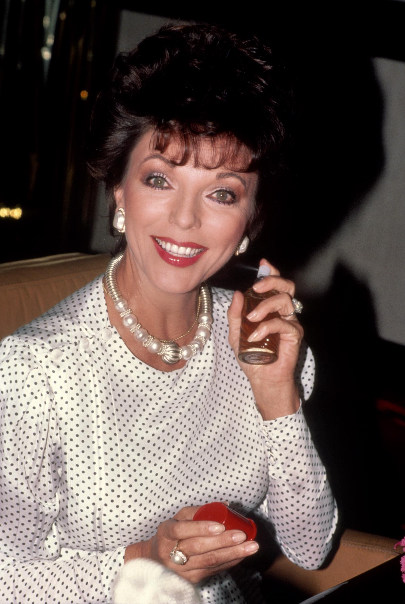 Joan Collins promotes Revlon's "Scoundrel" perfume in New York City. She wears a white polka-dot blouse with button detailing at the shoulder, accessorized with pearl and gold statement jewelry. Smiling brightly, she holds a perfume bottle in one hand and a red case in the other, showcasing her signature elegance.