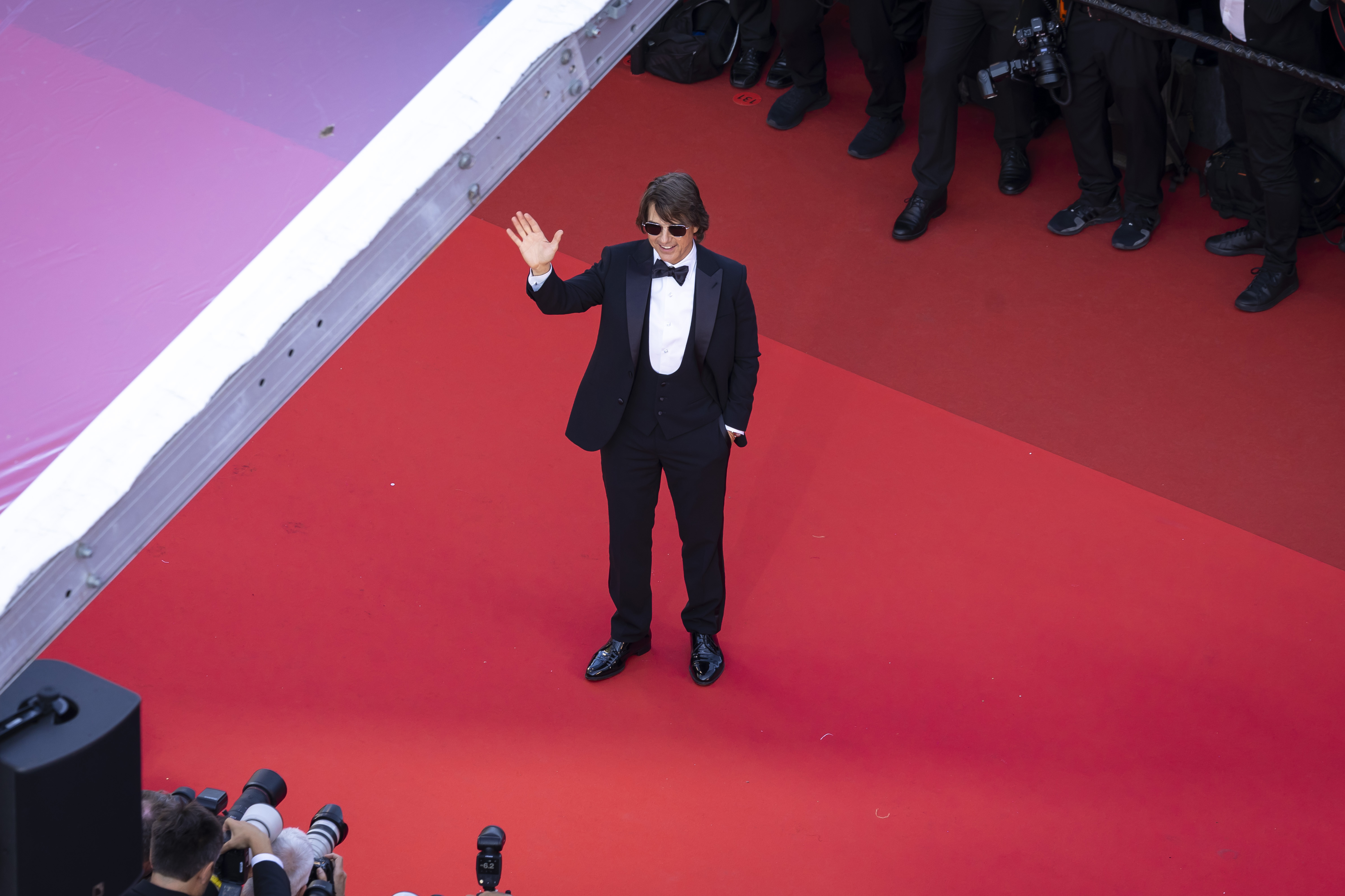 Seen from above, Tom Cruise arrives solo on the red carpet in a classic black tuxedo, offering a wave to the crowd during the "Mission: Impossible – The Final Reckoning" premiere at the 78th Cannes Film Festival.