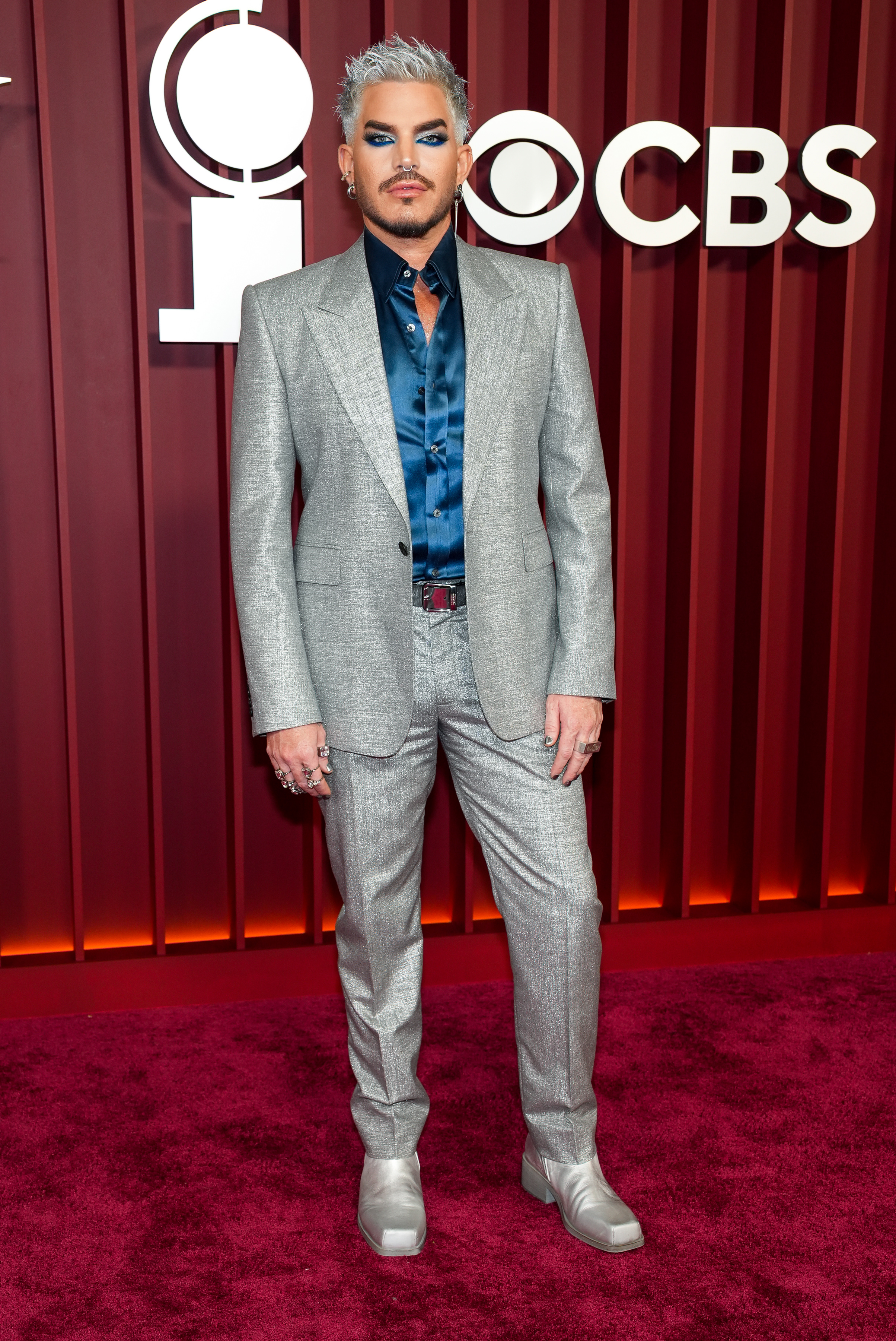 Adam Lambert stuns in a full-body shot, flaunting his shimmering suit and silver footwear. The deep blue silk shirt adds contrast, while his strong posture and sleek styling exude a blend of rock star bravado and red carpet elegance.