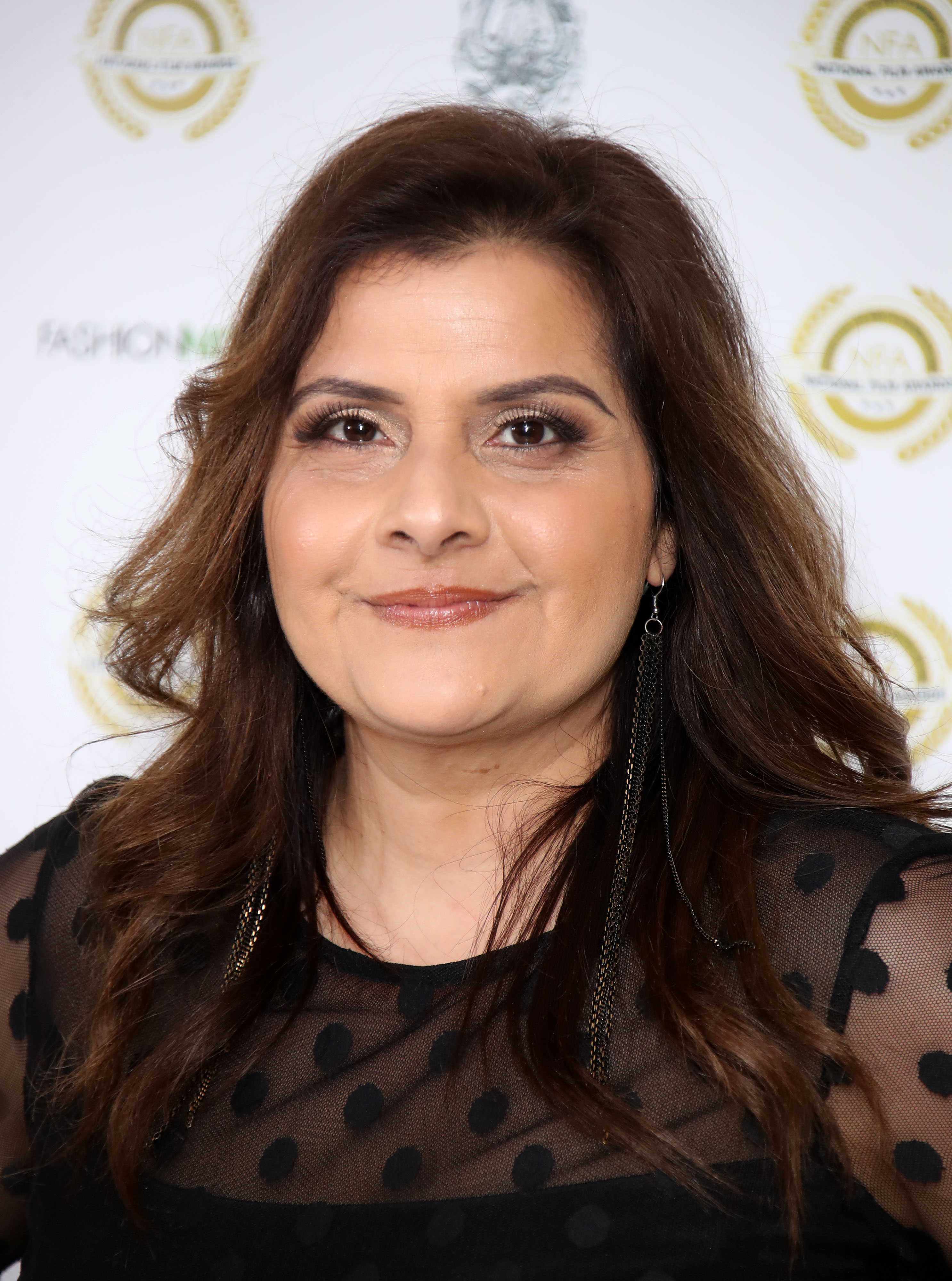 A close-up portrait of Wadia at the National Film Awards at Porchester Hall in March 2019, her dark hair now lightened with warm brown tones and styled in long loose waves. She wears a sheer black polka-dot top and long chain tassel earrings, her eyes framed with a smoky bronze shadow.