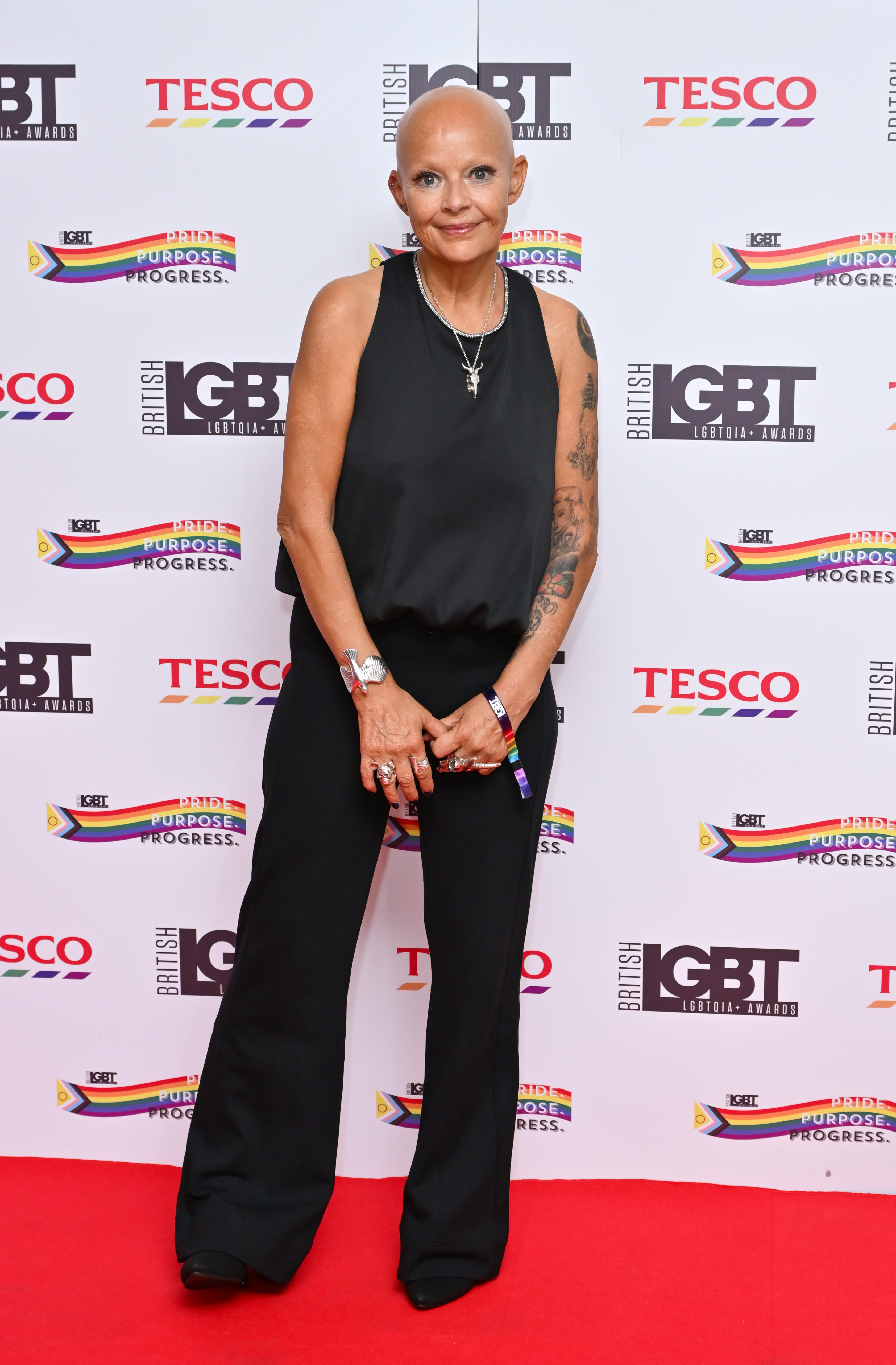 Porter stands full-length on a red carpet in a sleeveless black satin top and wide-leg trousers, her arms — covered in extensive tattoo work from shoulder to wrist — left completely bare. Multiple rings stack on her fingers and a rainbow wristband sits at her wrist, the British LGBT Awards backdrop stretching behind her. Pictured at the British LGBT Awards at The Brewery, London, June 2025.