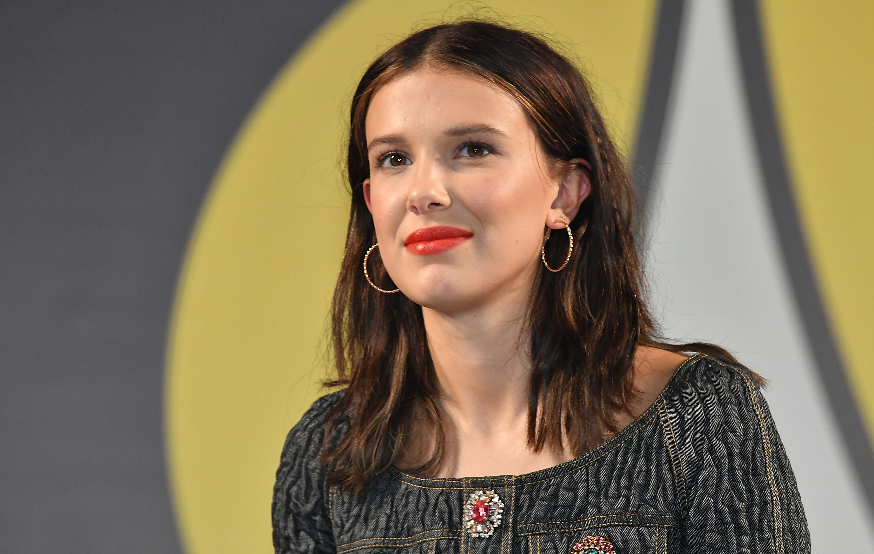 Millie Bobby Brown appears youthful yet assured with softly styled hair and gold hoop earrings. A bold red lip adds contrast to the understated outfit. The look captures a transitional moment between girlhood charm and rising star presence.