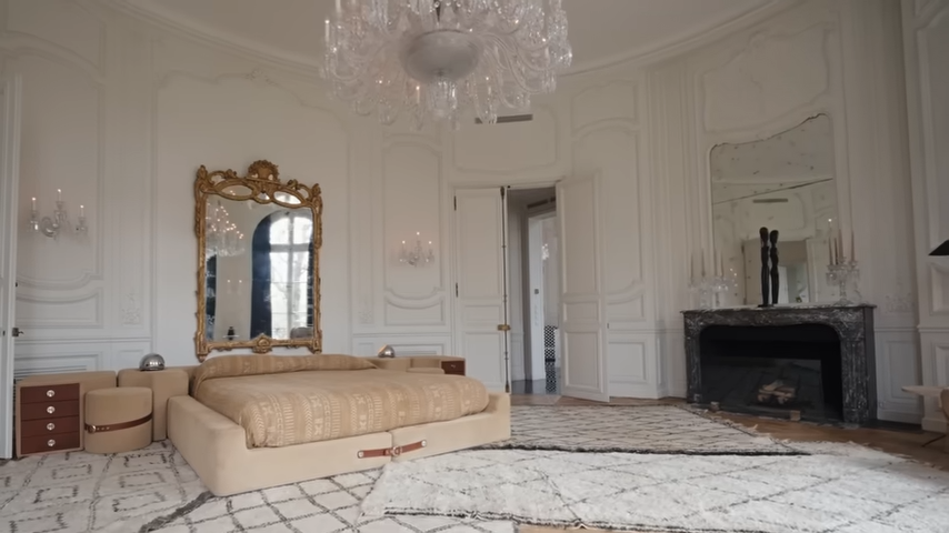 The bed is perfectly positioned beneath a towering, ornate gold-gilded mirror and flanked by a classic black stone fireplace. | Source: YouTube/@Archdigest