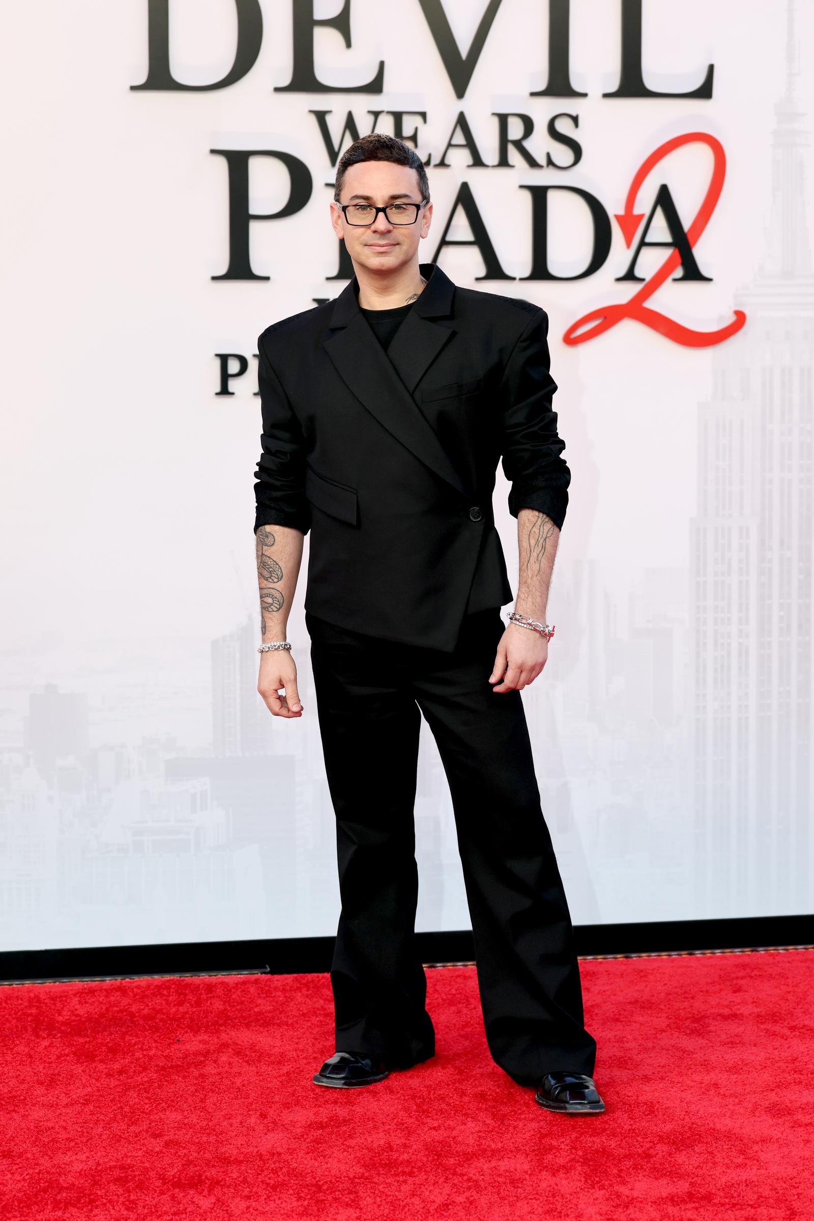 Christian Siriano goes head-to-toe black in a sleeveless double-breasted blazer over a black T-shirt and wide-leg trousers, adding a flash of silver with diamond bracelets on both wrists, black-framed glasses, and black loafers.