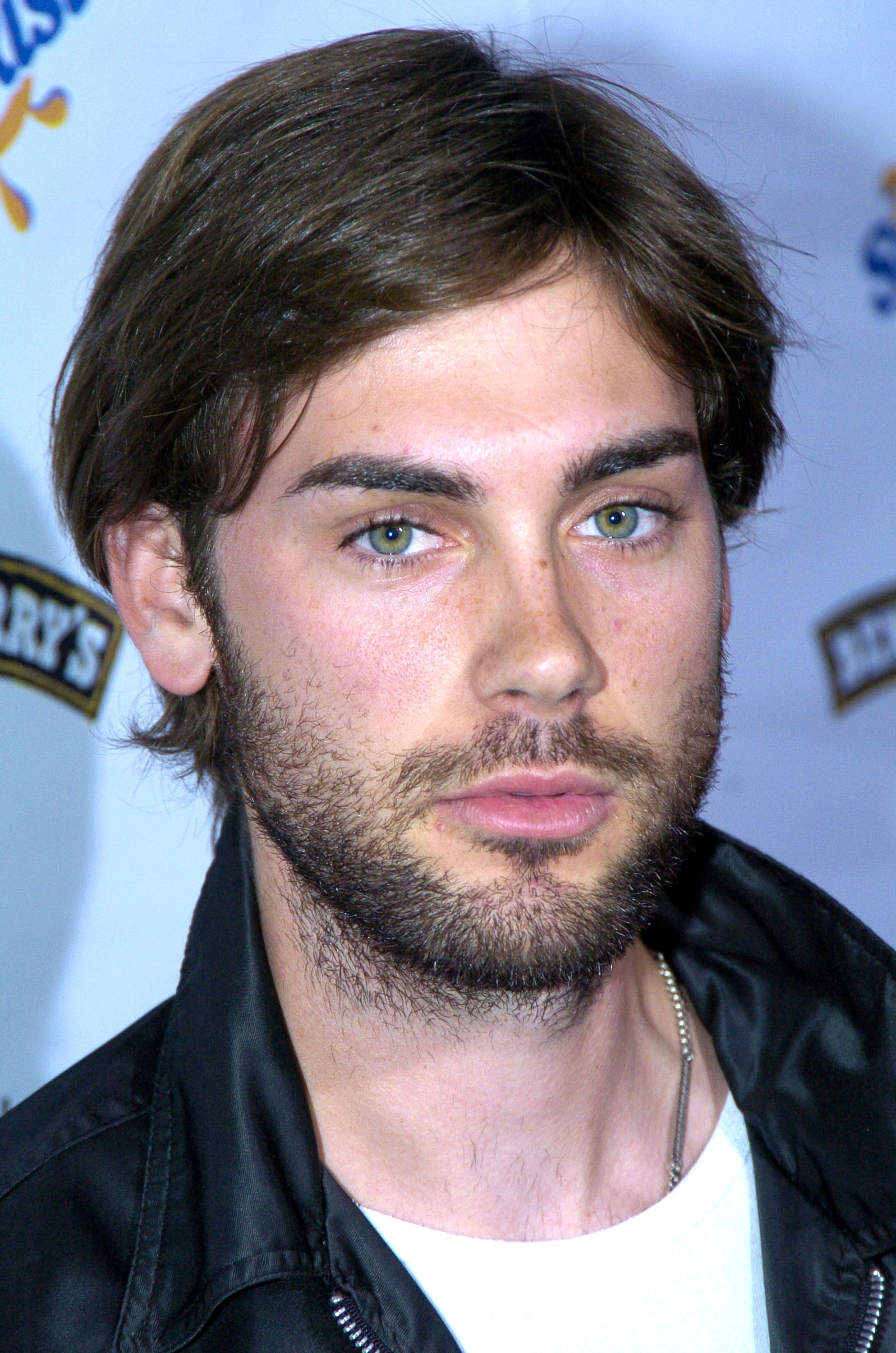 Drew Fuller appears at a public event in the mid-2000s, sporting a more rugged look with facial hair and tousled hair. Dressed in a black leather jacket over a white T-shirt, he gazes into the camera with a quiet intensity. The casual yet edgy styling reflects a shift from his earlier clean-cut image.